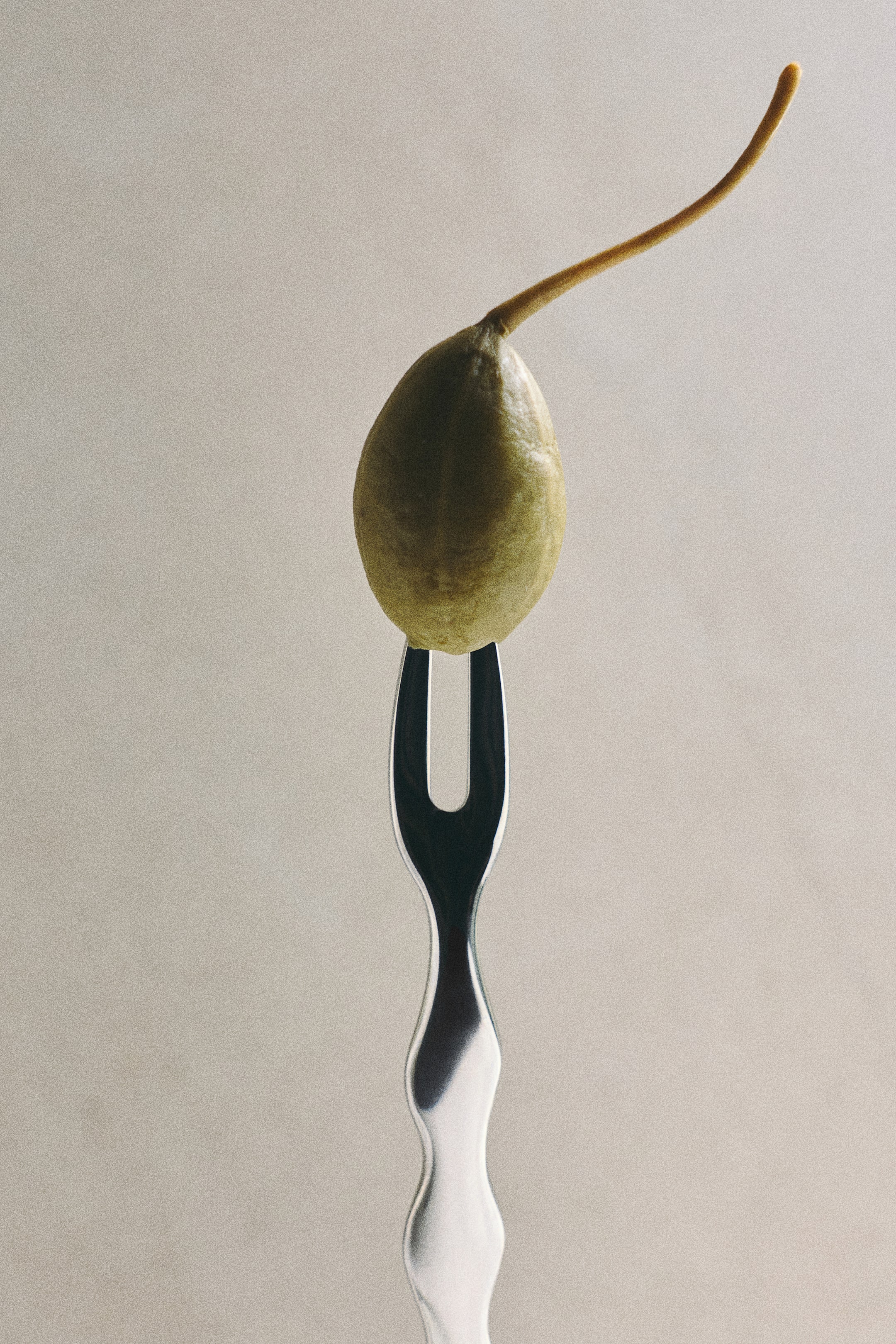 View larger image: A small fork in polished stainless steel featuring a wavy, contoured handle is holding a single green caper berry on its two tines, shown from a front close-up view.