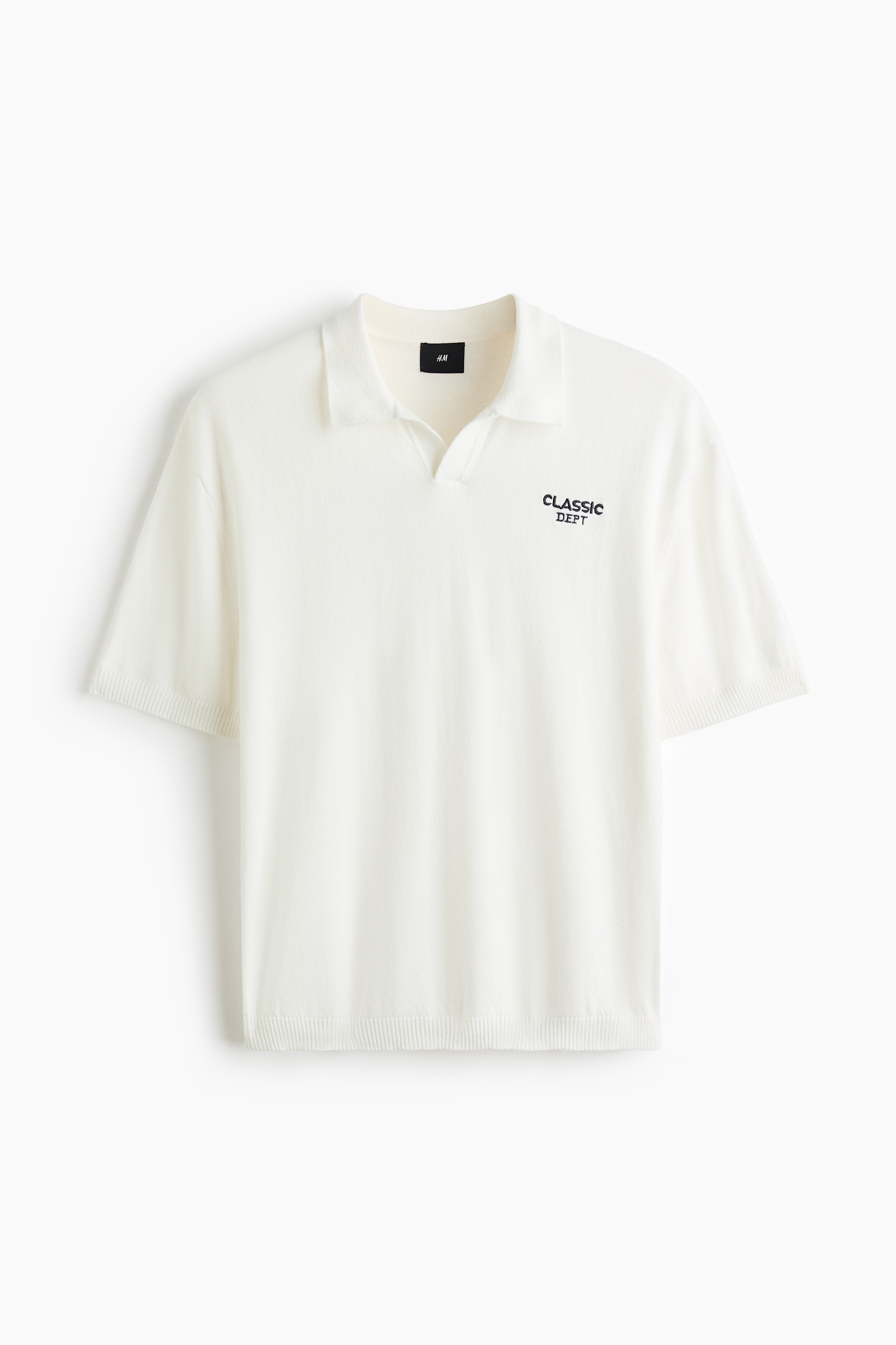 A short-sleeved, off-white knit polo shirt is being worn by the model, featuring a collar, ribbed cuffs, a ribbed hem, and a small black embroidered text motif on the chest. The loose-fitting polo is paired with light-coloured trousers while the model is sitting.