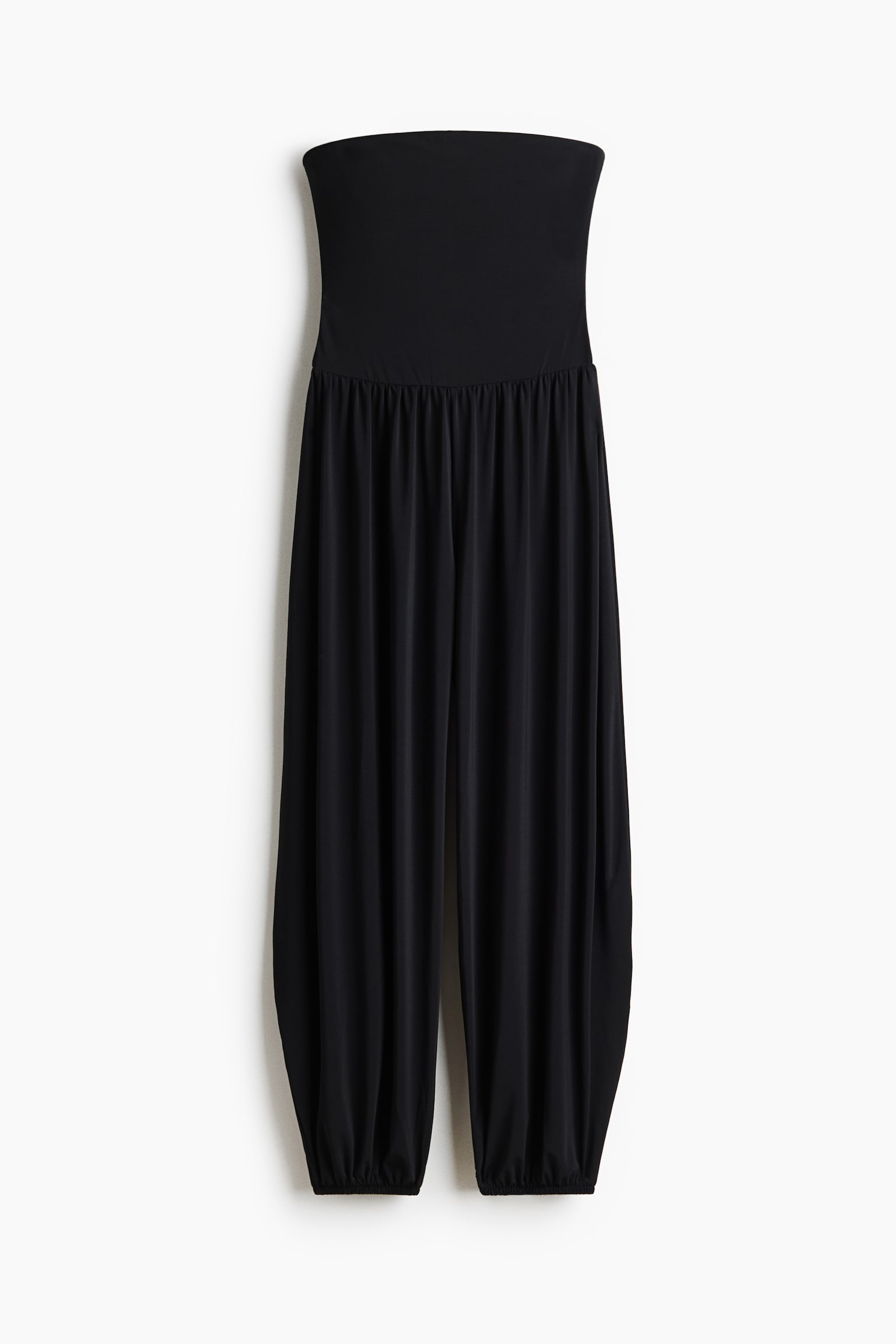 A black strapless jumpsuit features a fitted bandeau bodice. The waistline showcases elasticated gathers, while the full, flowing legs are cinched at the ankles with elasticated cuffs.