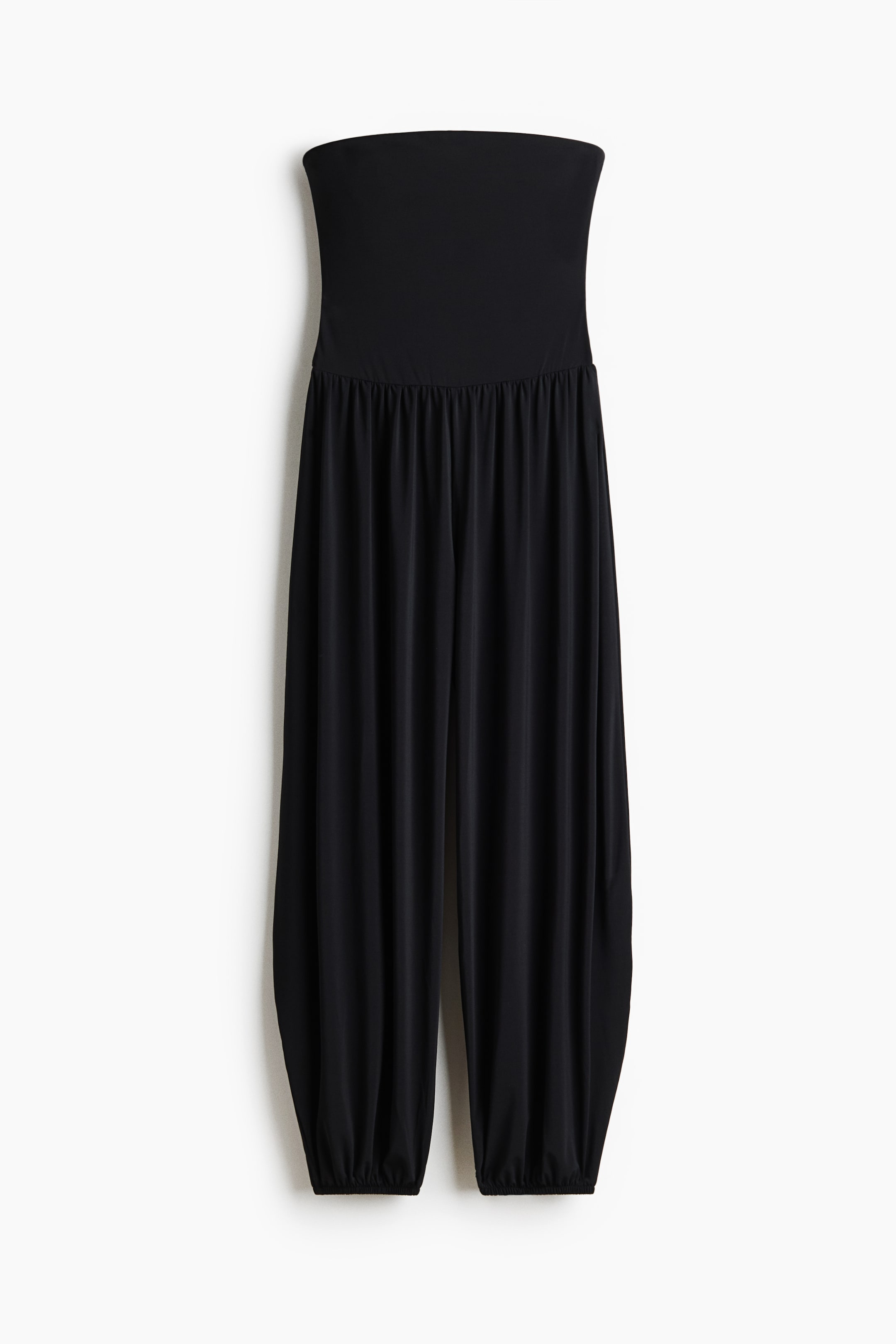 View larger image: A black strapless jumpsuit showcasing a fitted bandeau bodice. The waistline highlights elasticated gathers, and the full, flowing legs taper at the ankles with elasticated cuffs.
