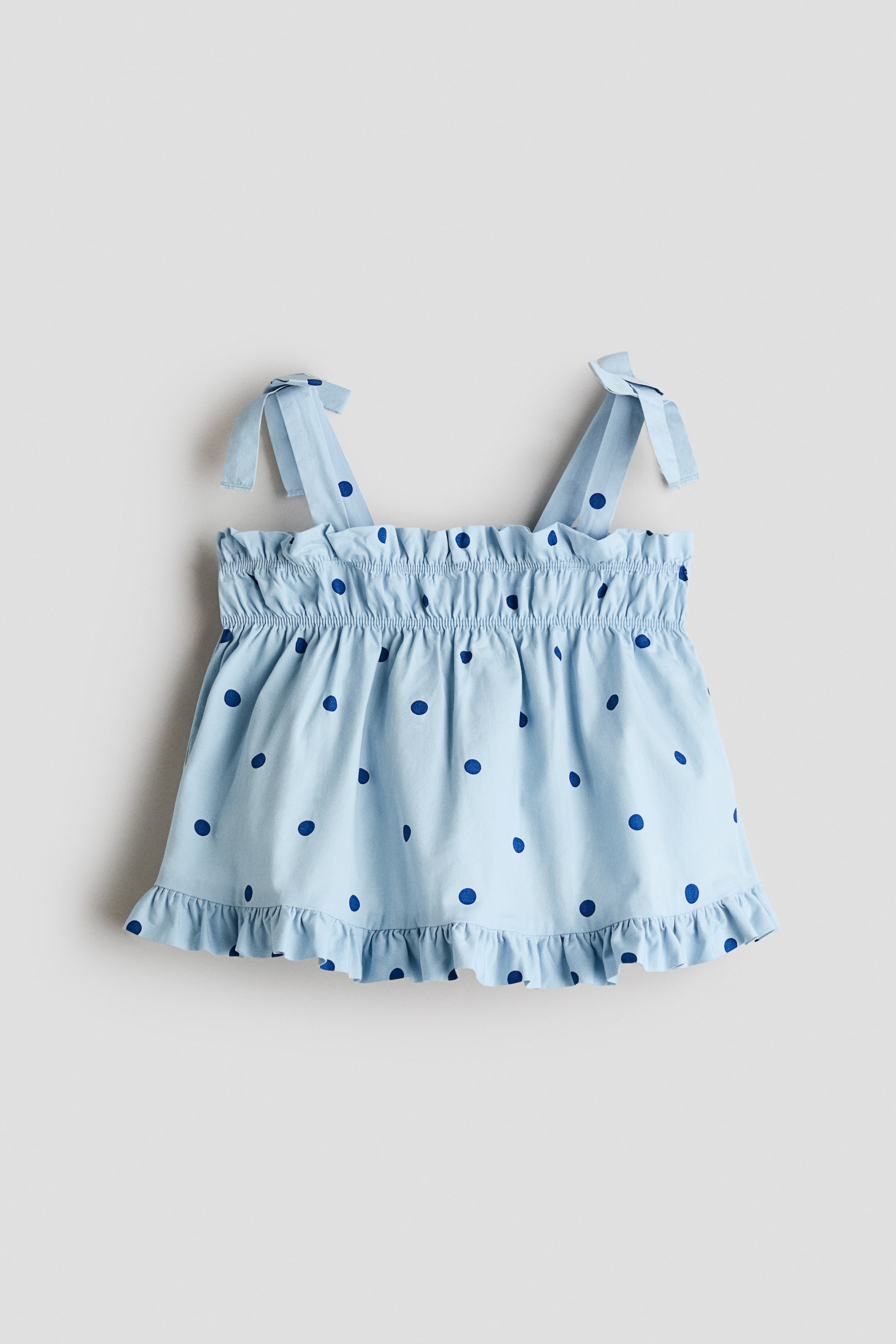 View larger image: The light blue sleeveless top is facing front, displaying a dark blue polka dot pattern and thick shoulder straps tied in bows. It features a smocked, elasticised chest with a ruffled upper trim and a flared, gathered body ending in a ruffled hem.