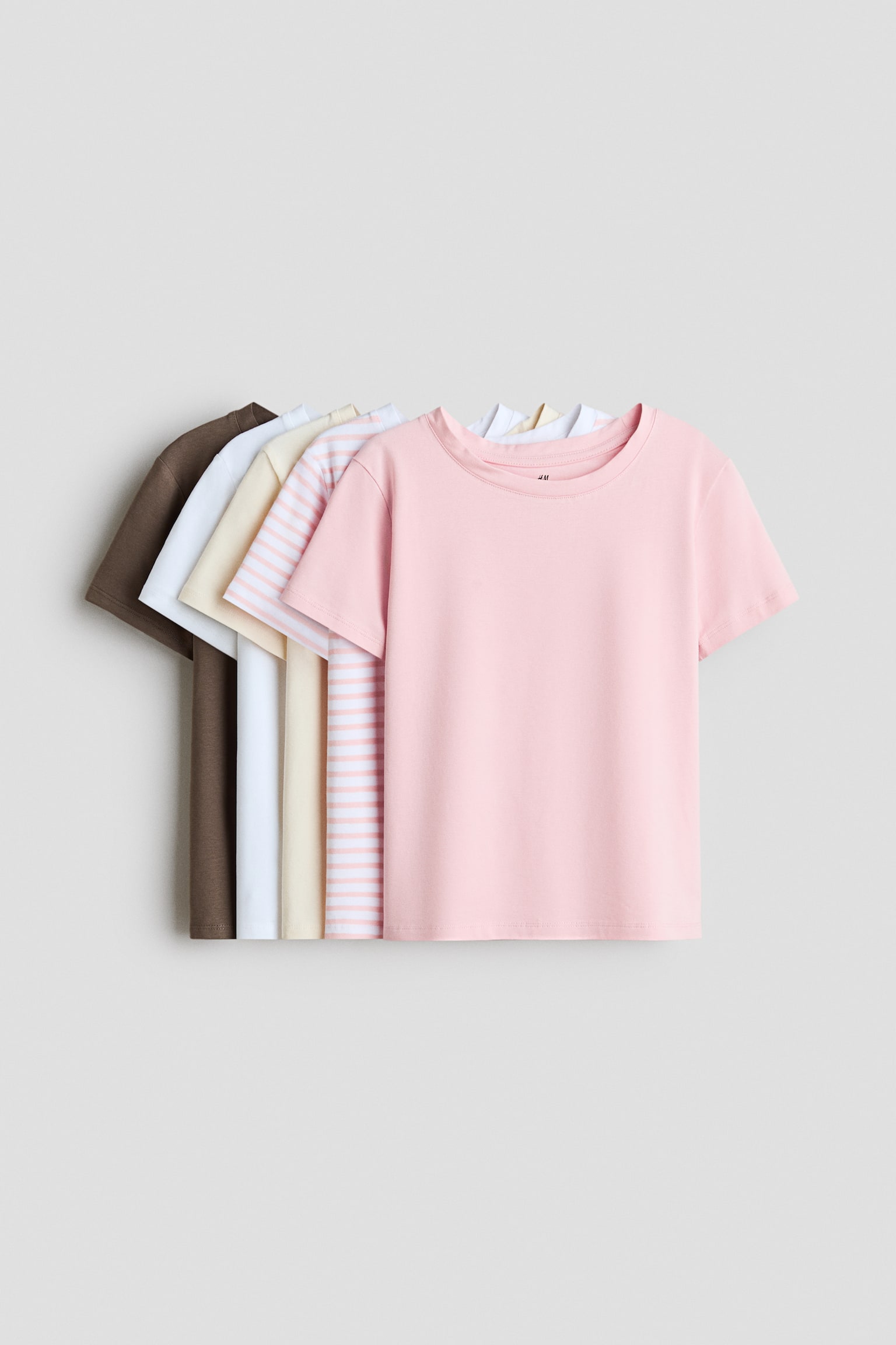 A set of five crewneck, short-sleeved t-shirts is being presented in a layered arrangement, with the front of a solid light pink t-shirt fully visible. The remaining shirts are fanning out to the left, showing pink-and-white striped, cream, white, and dark brown colorways.