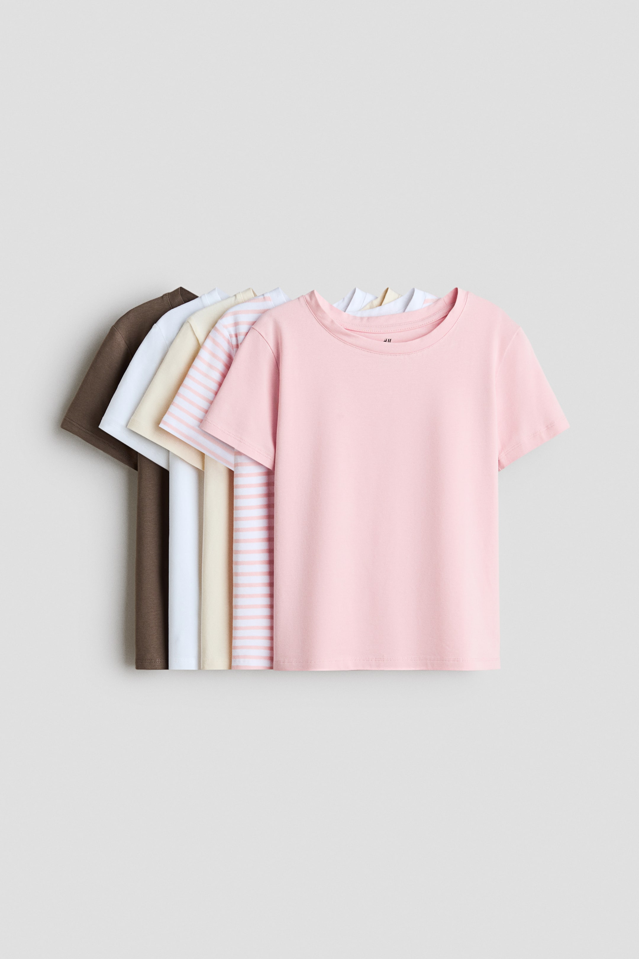 View larger image: A set of five crewneck, short-sleeve T-shirts is displayed in a layered arrangement, with the front of a solid light pink T-shirt fully visible. The remaining shirts fan out to the left, showing pink-and-white striped, cream, white, and dark brown colorways.