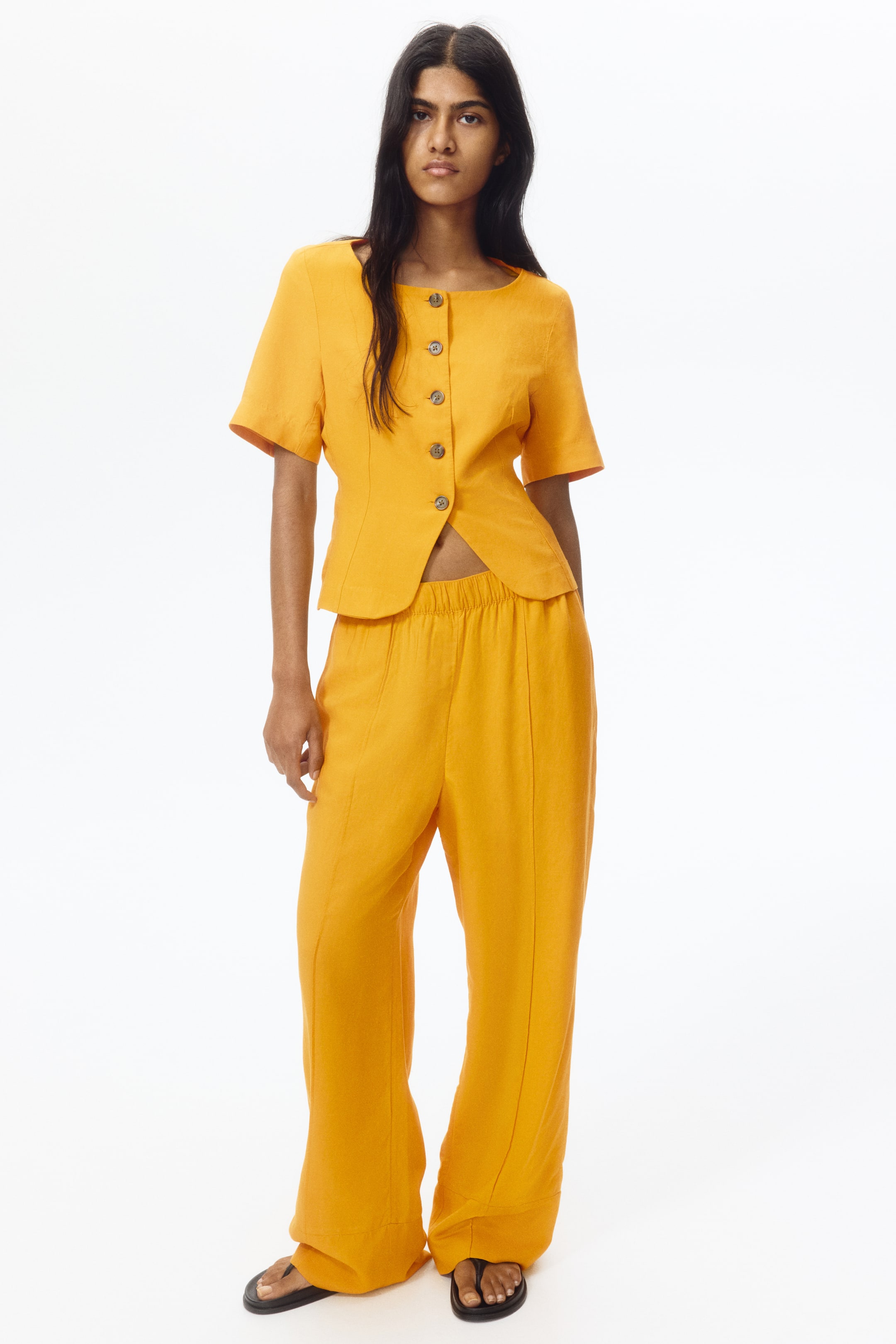 View larger image: Introducing an orange textured two-piece set, comprising a short-sleeved top with a round neckline, front button closure, and a shaped hem. The matching wide-leg pants come with an elasticated waist and vertical seam details.
