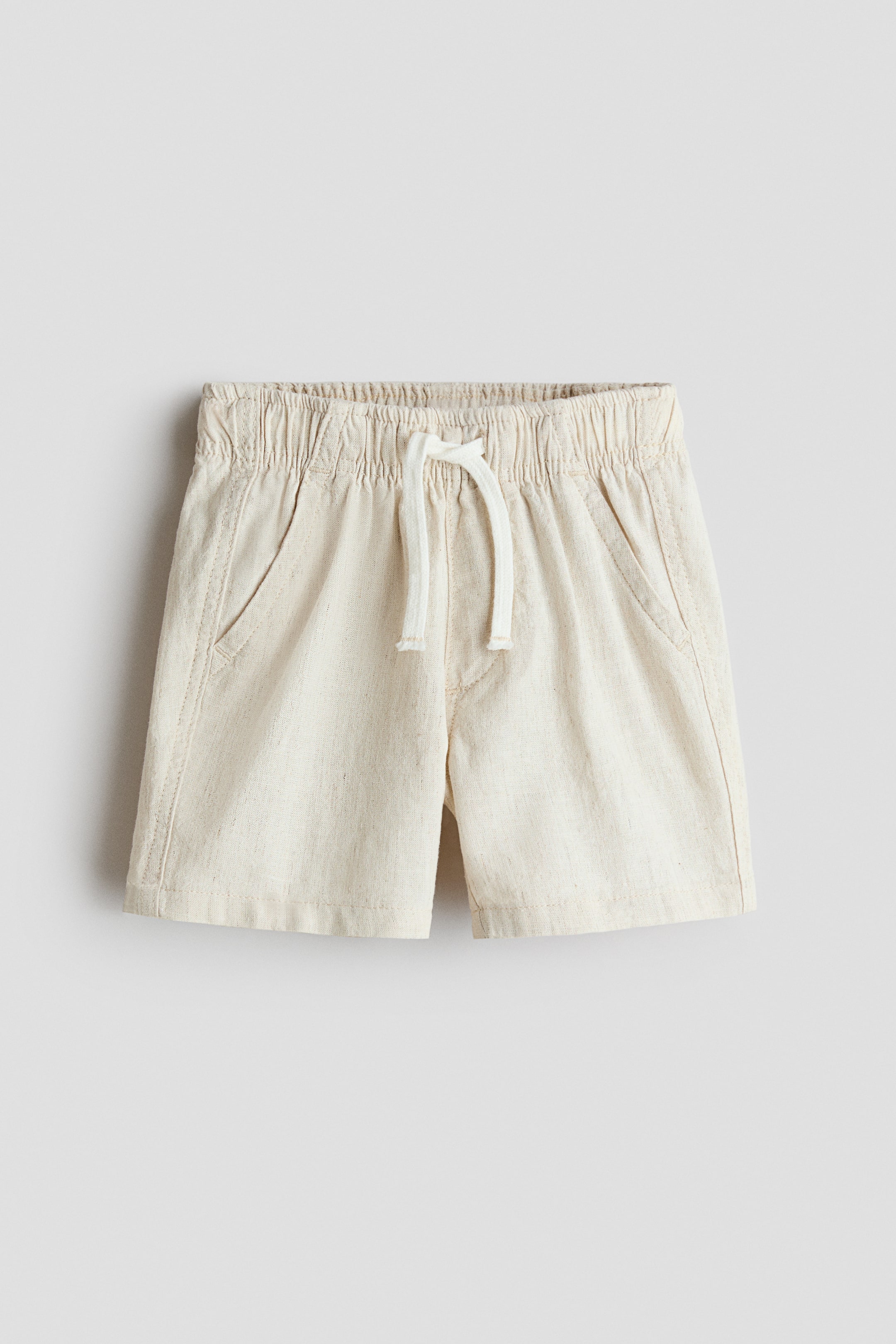 View larger image: A pair of light-colored shorts is displayed from the front, showcasing a soft, textured weave, an elasticated waistband with a drawstring and light-colored tie, decorative front pockets, and a stitched detail indicating a faux fly.