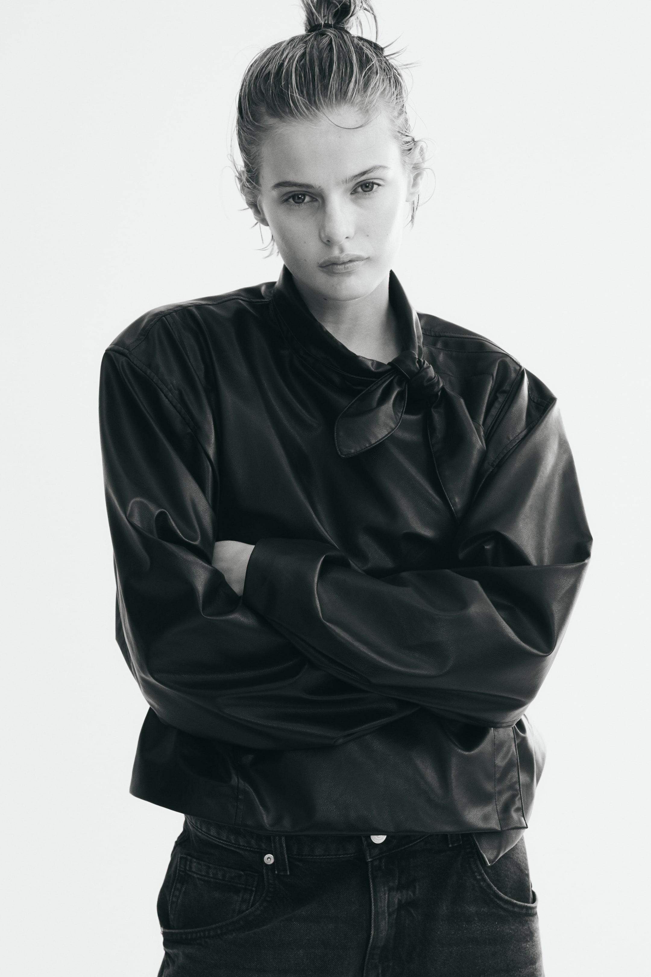 View larger image: An oversized, short, dark-colored jacket in coated fabric, featuring dropped shoulders and a scarf collar tied in a knot at the front, is being worn by the model. The model is crossing their arms across the jacket, which is styled with dark-colored jeans.