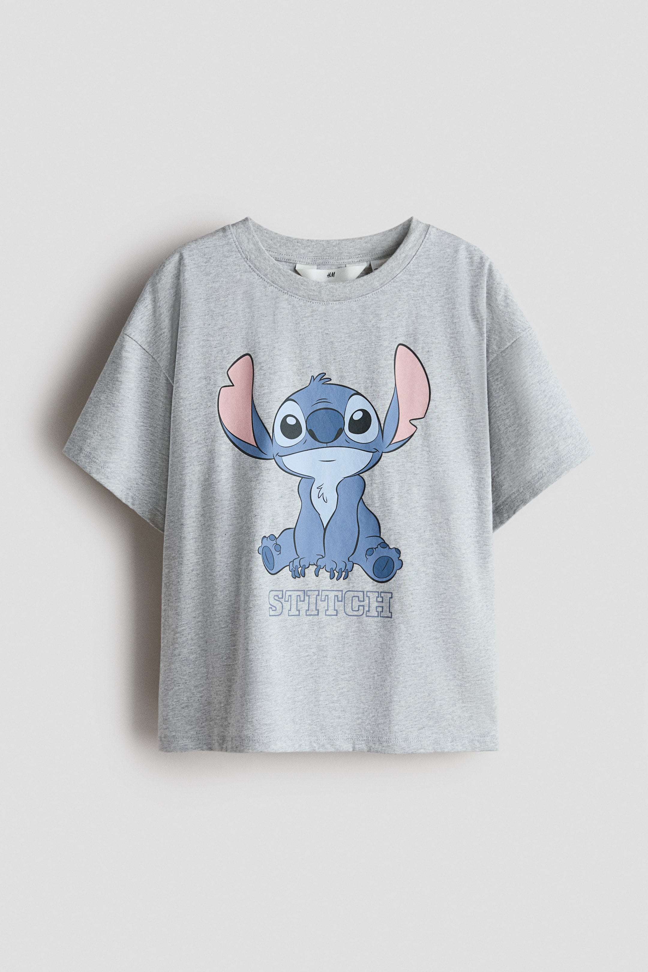 View larger image: A gray oversized t-shirt in soft, printed cotton jersey is presented FRONT, featuring a round, rib-trimmed neckline and dropped shoulders. The front is displaying a large blue and pink graphic of the character Stitch sitting, with the word "STITCH" printed below.