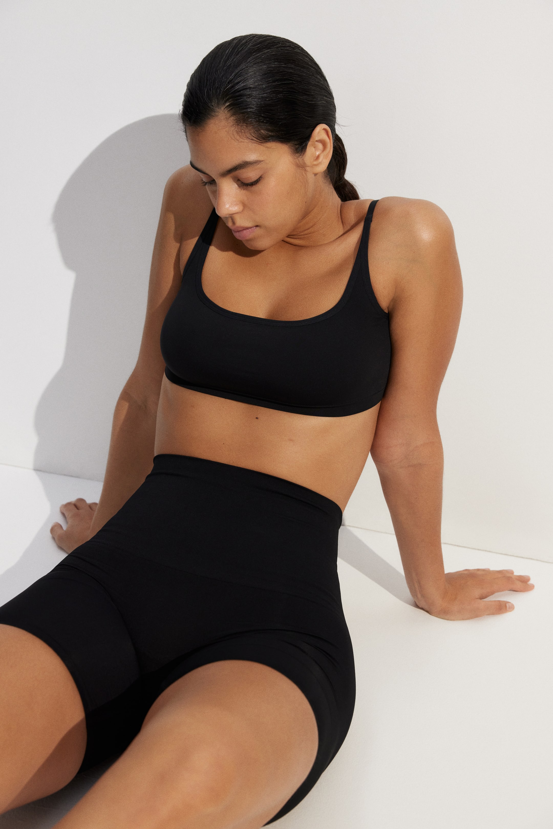 Se større billede: En sort sports-bh med dyb rund halsudskæring og brede stropper bæres af modellen, kombineret med sorte, højtaljede cykelshorts med sømløst design.