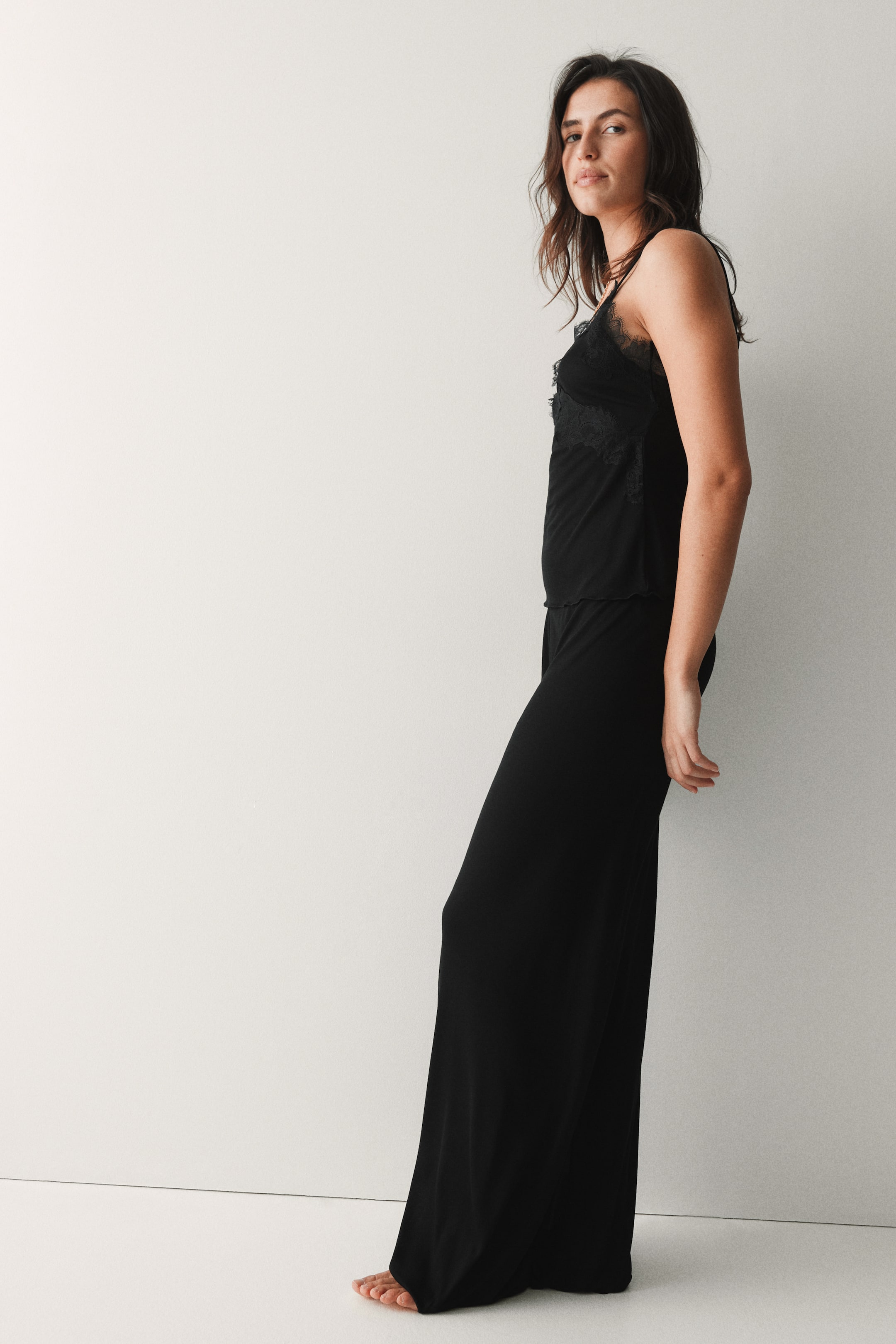 Voir une image plus grande: Un ensemble de pyjama noir deux pièces comprenant un haut camisole à fines bretelles avec des détails en dentelle noire le long de l'encolure, et un pantalon ample à jambes larges tombant jusqu'au sol.