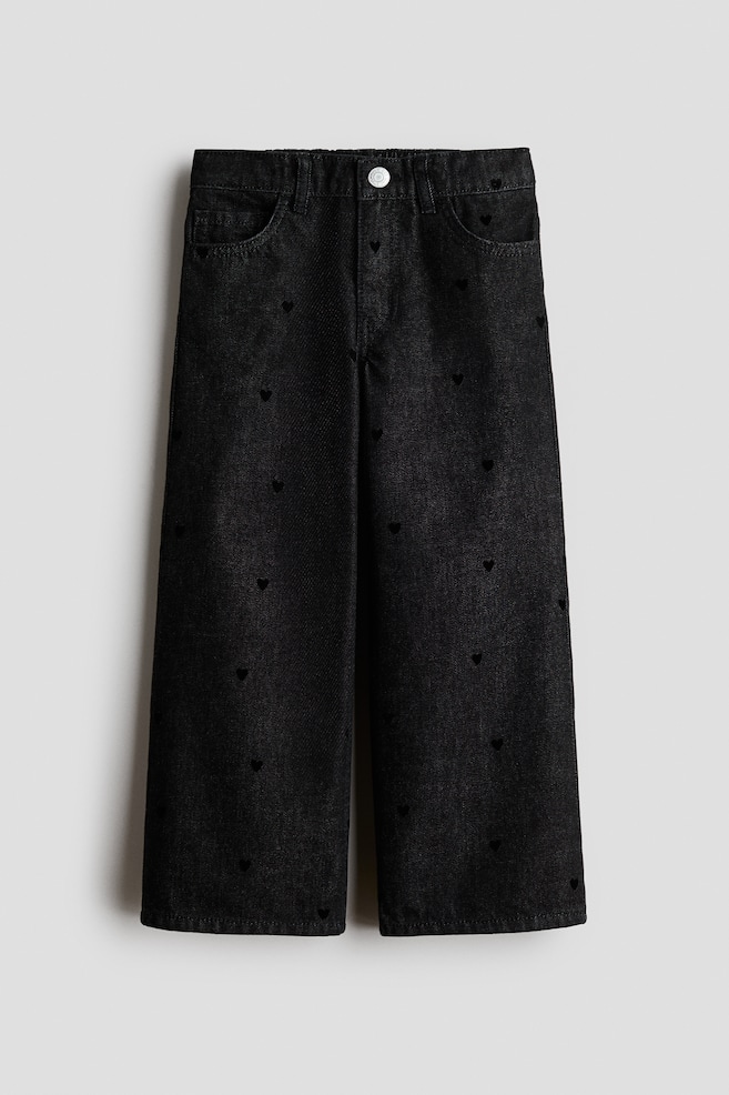 Une paire de jeans en denim de coton noir est présentée de face, avec une coupe ample, des jambes larges et un motif intégral de petits cÅurs noirs brodés. Le jean présente un design classique à cinq poches, des passants de ceinture et une taille avec une seule pression métallique. - 1