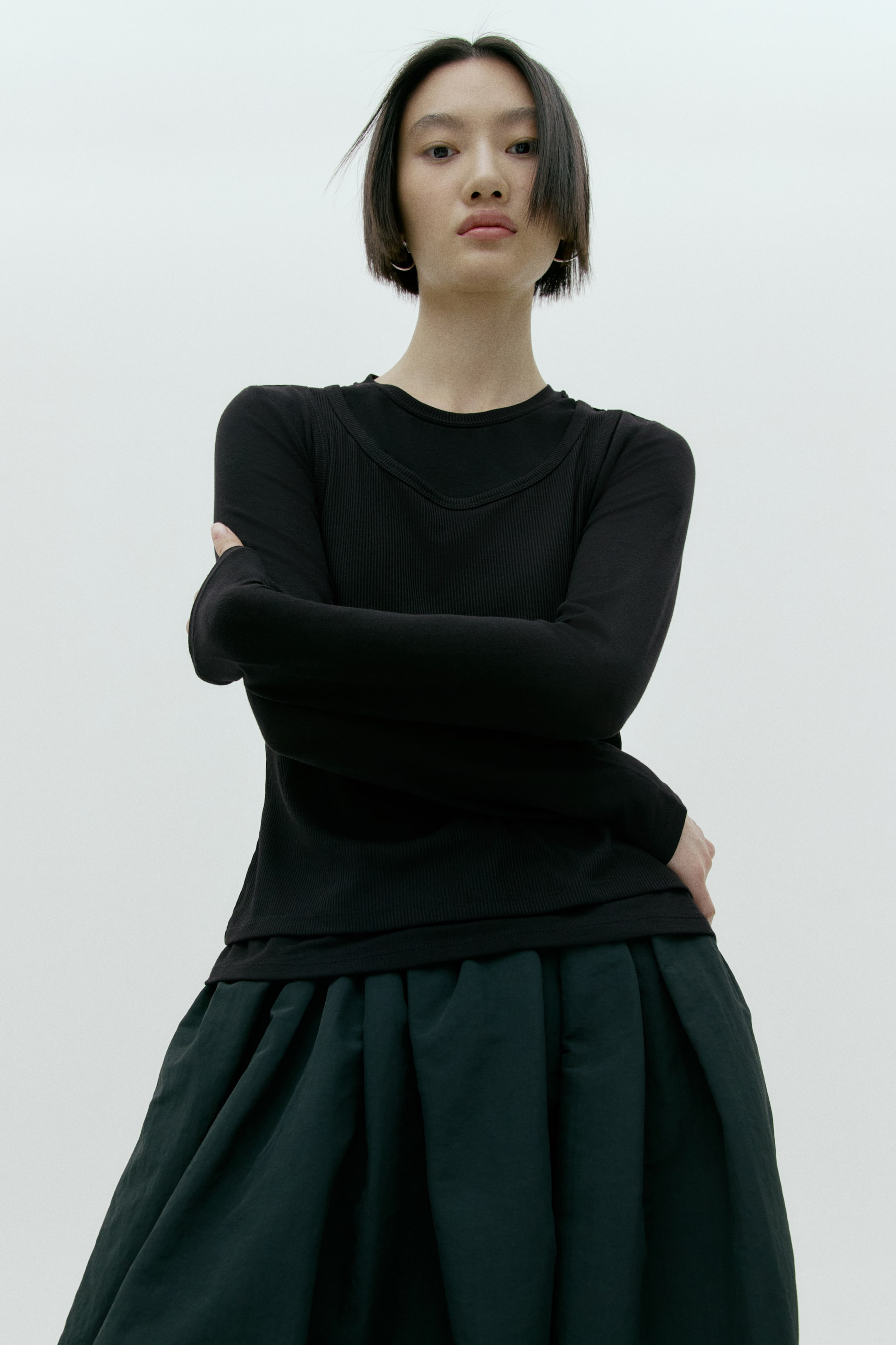 拡大画像を表示: 黒の長袖トップはレイヤードデザインで、滑らかなラウンドネックの下にリブのVネックパネルを重ねている。ダークグリーンのフルスカートは深いプリーツが施され、ボリュームのあるシルエットを作り出している。アンサンブルはやや低めの正面アングルから撮影されている。