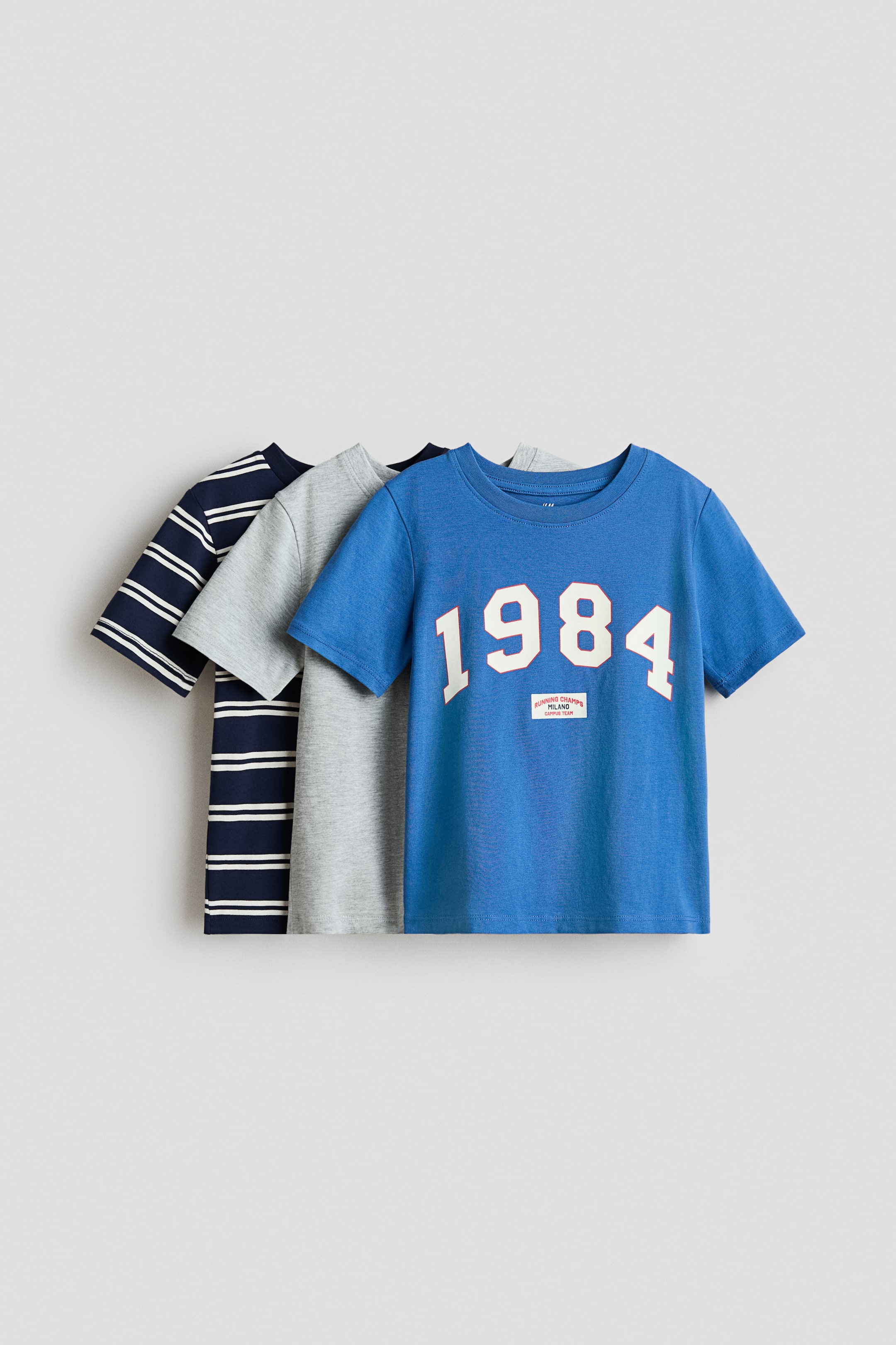 View larger image: Three classic t-shirts in soft cotton jersey are shown layered, each featuring round necklines and short sleeves. The front blue t-shirt displays a white "1984" graphic on its front, while the middle t-shirt is solid light gray, and the back one is navy blue with white horizontal stripes.