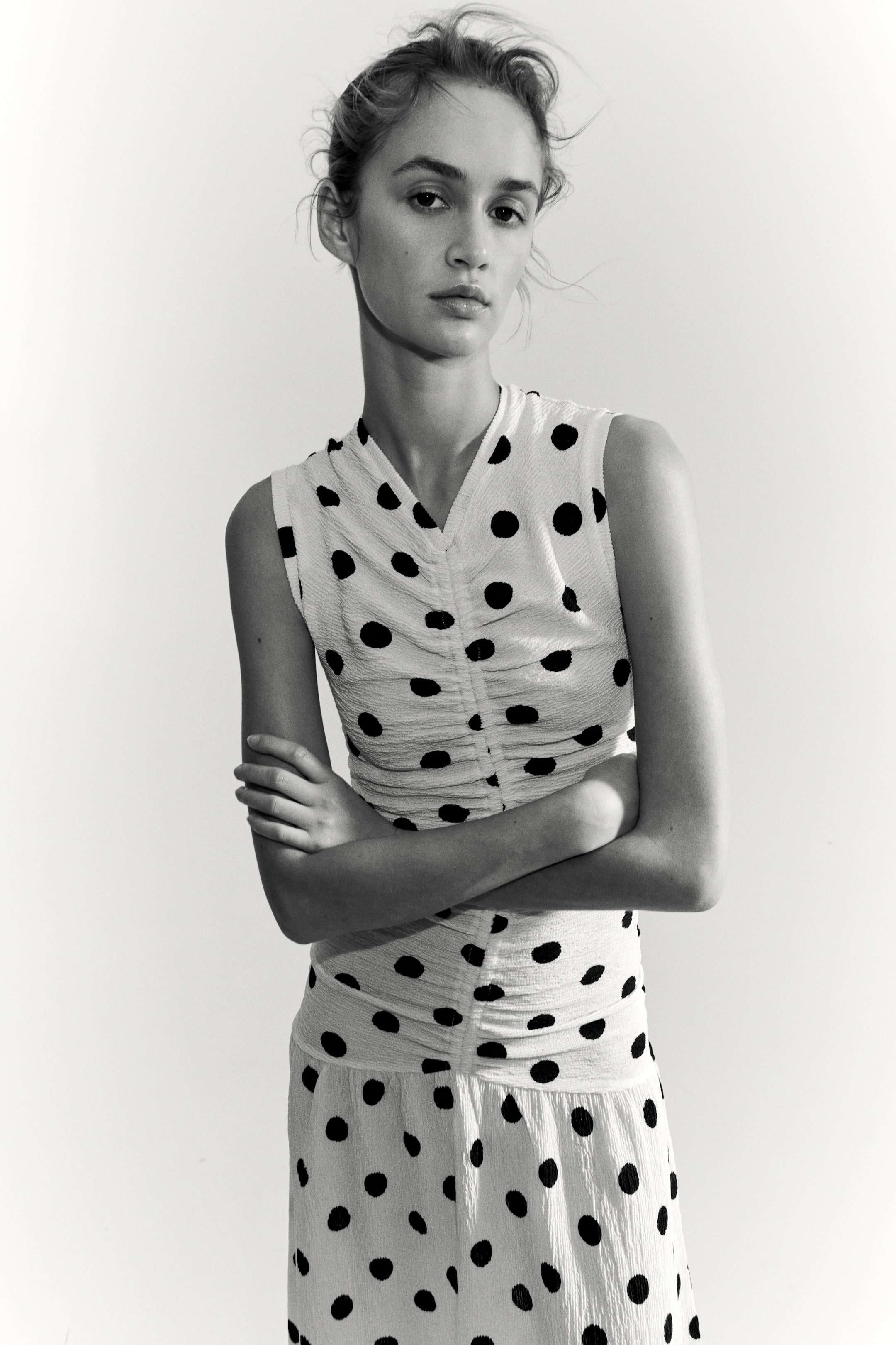 View larger image: The model is wearing a long, sleeveless dress crafted from light-coloured crinkled jersey adorned with dark polka dots. This dress boasts a V-neckline and features an elasticated seam at the front, creating gathers, along with a gathered seam below the hips that transitions into a flared skirt. The model is posed with her arms crossed.