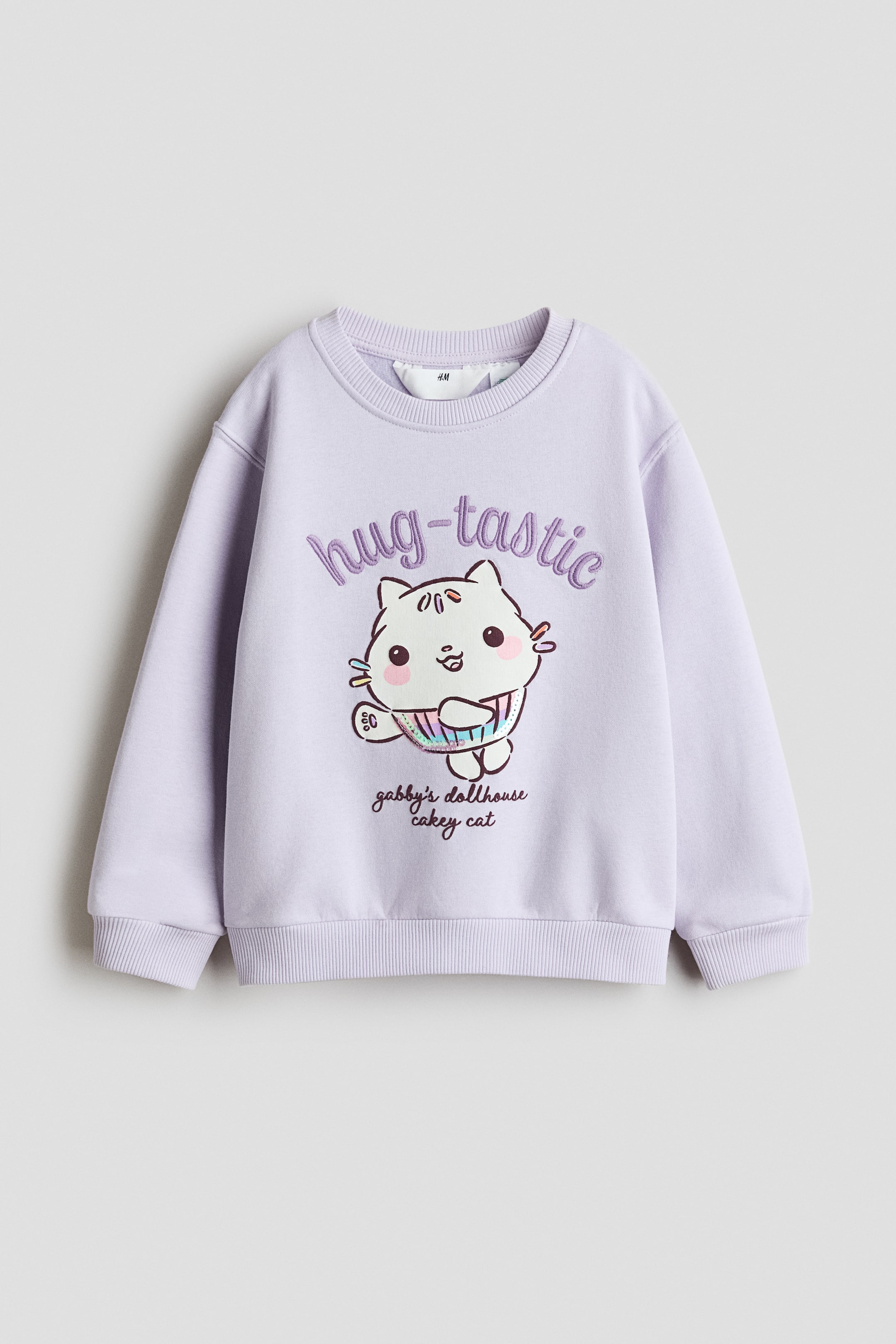 View larger image: A front-facing lavender sweatshirt features a ribbed crew neck, ribbed cuffs and a ribbed hem. The front displays a graphic of a cartoon white cat wearing a rainbow cupcake wrapper, with the embroidered text "hug-tastic" above and "gabby's dollhouse cakey cat" below.