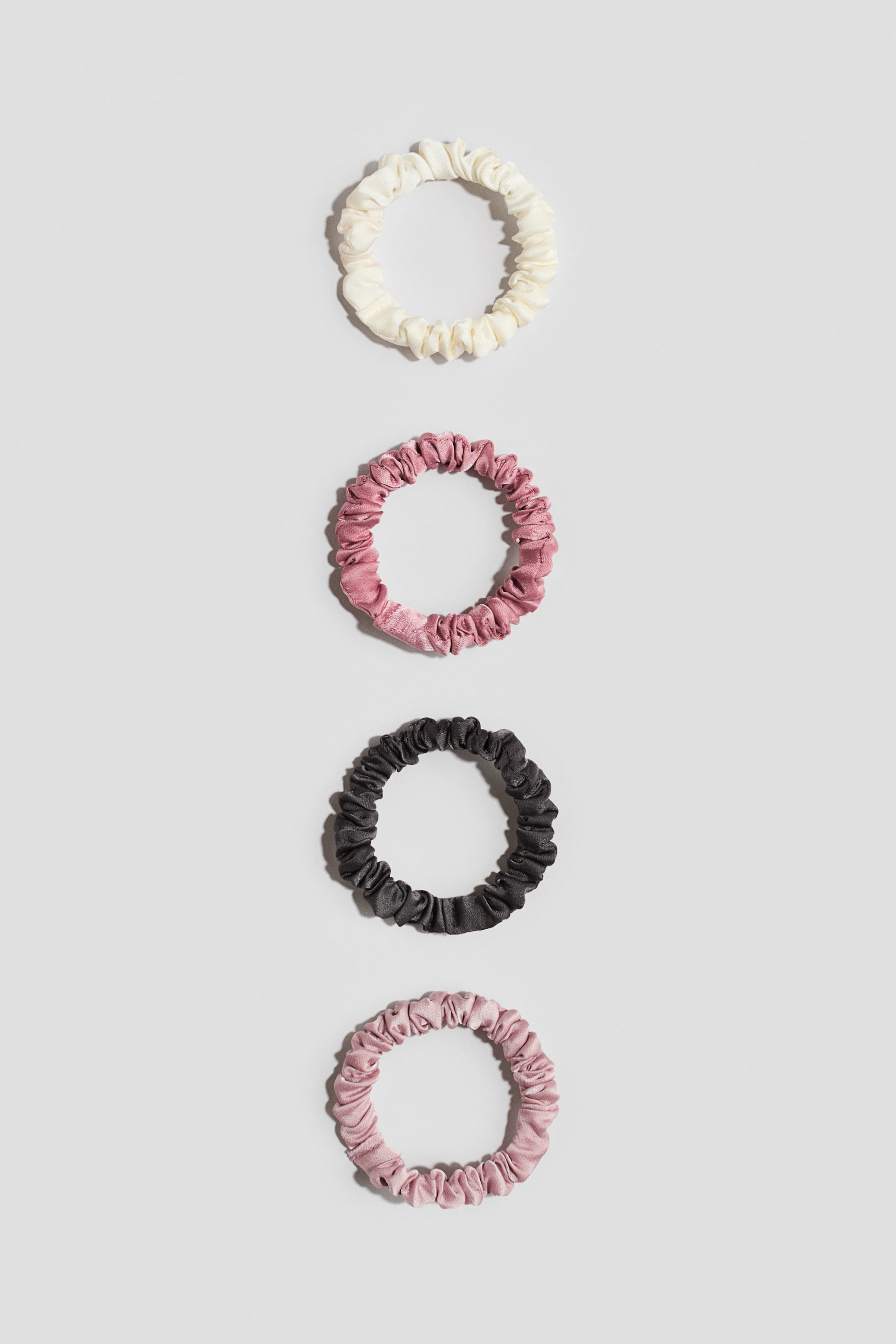 View larger image: Four scrunchies are arranged vertically, showing a gathered texture. They are made of woven fabric with a slight sheen and are presented in off-white, dusty rose, dark grey, and light pink colours.