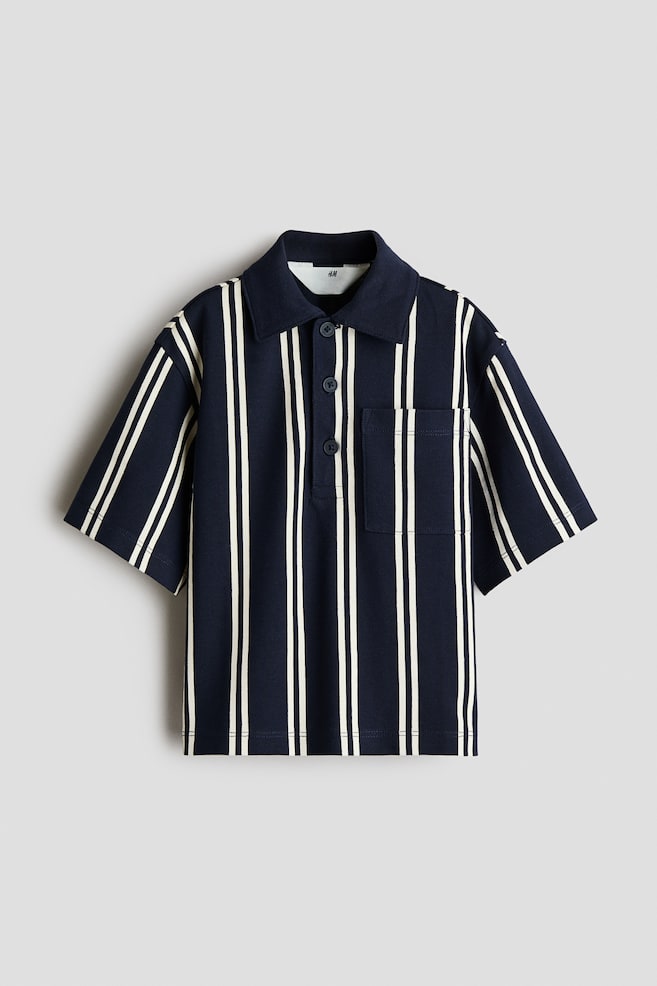 Una camiseta polo de manga corta azul marino presenta rayas verticales blancas, cuello con solapas, una tapeta de tres botones y un bolsillo de parche en el pecho izquierdo. - 1