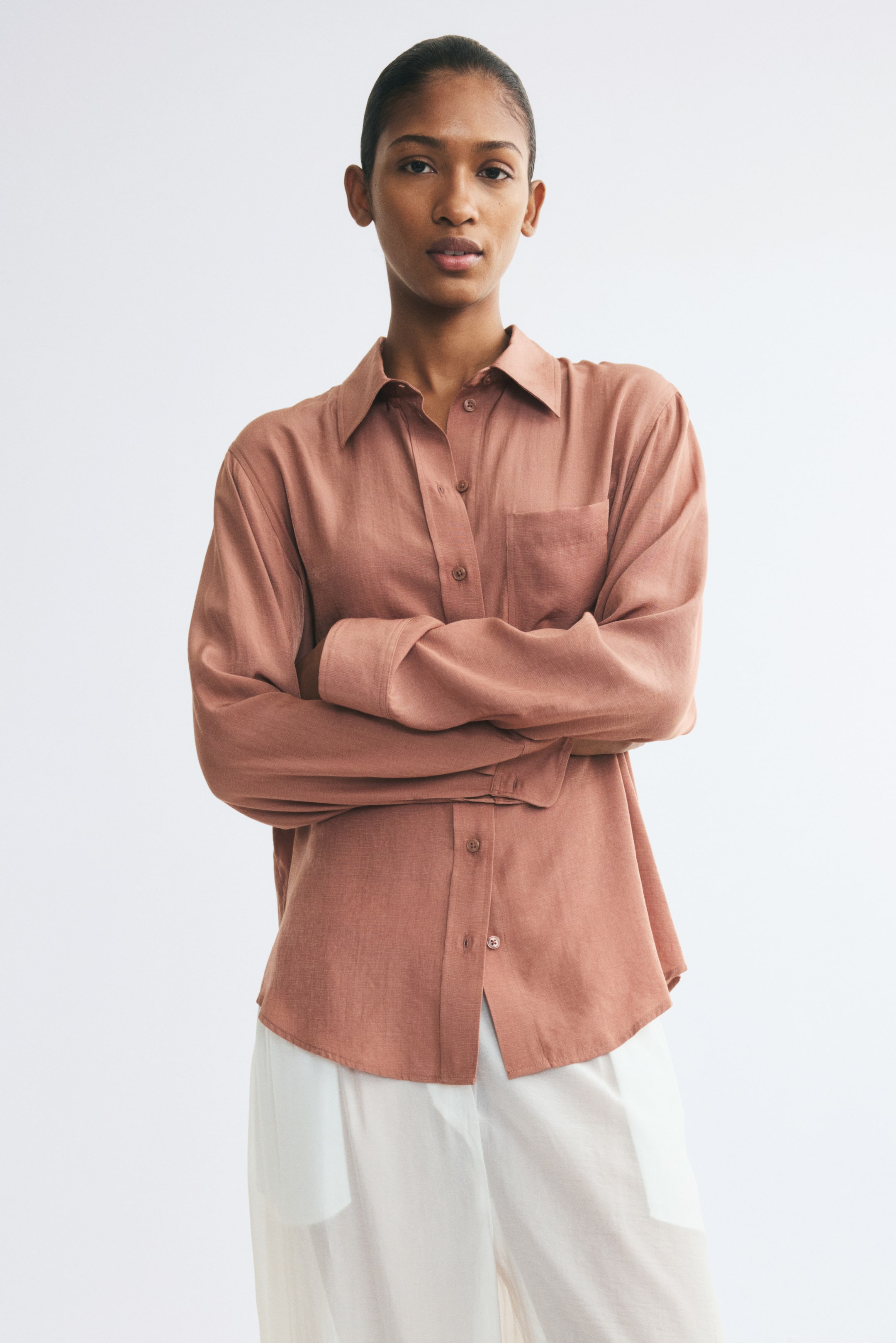 View larger image: A terracotta-colored shirt in a soft weave is worn by the model with arms crossed. The shirt features a collar, a single patch chest pocket, long sleeves with button cuffs, and a rounded hem, styled with white trousers.