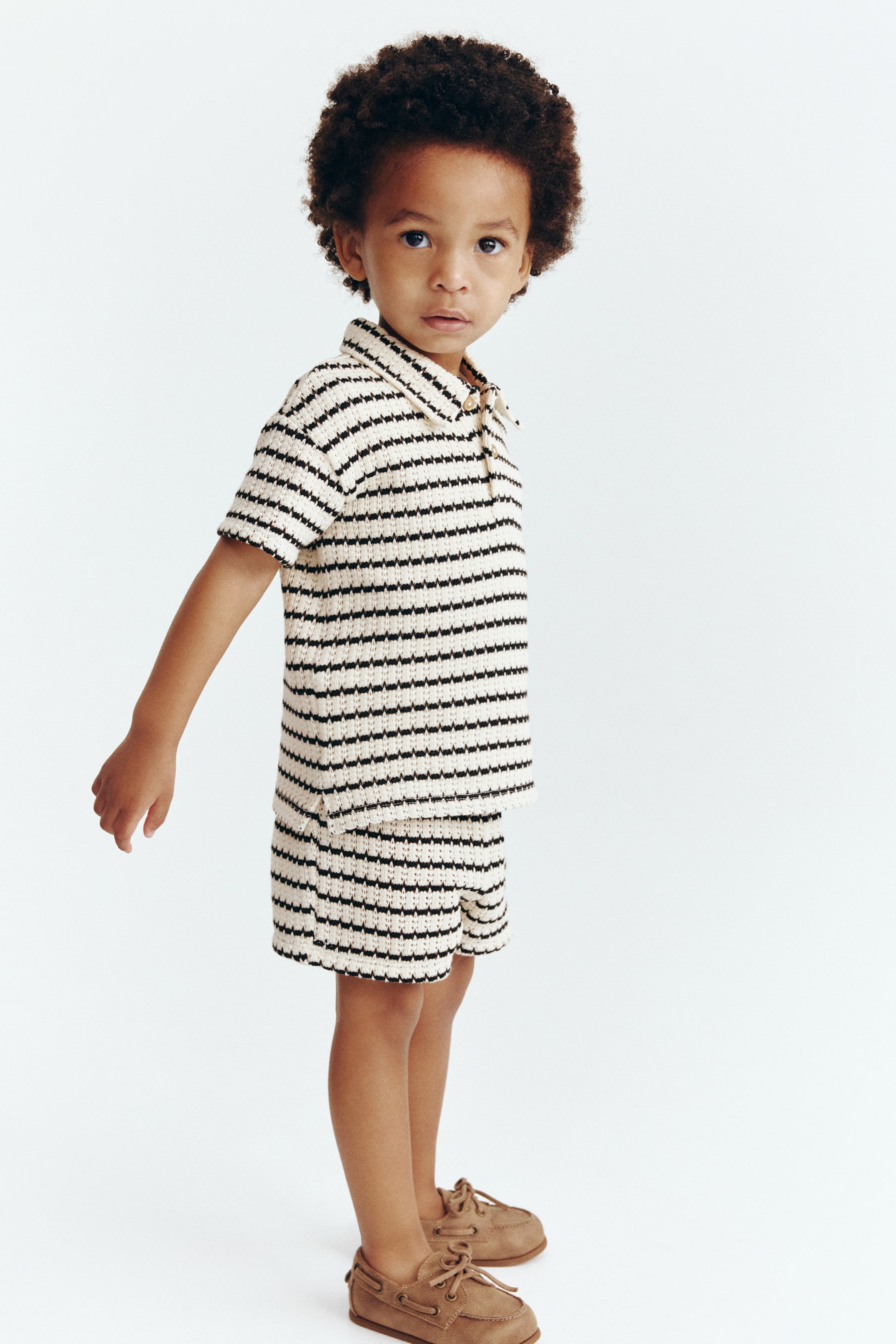 Ampliar la imagen: Una camiseta polo de punto con rayas horizontales en crema y negro y shorts a juego se presentan desde un ángulo ligeramente lateral. La camiseta polo cuenta con un cuello, mangas cortas y una tapeta de dos botones, mientras que los shorts muestran un bolsillo lateral y aberturas laterales en el dobladillo.