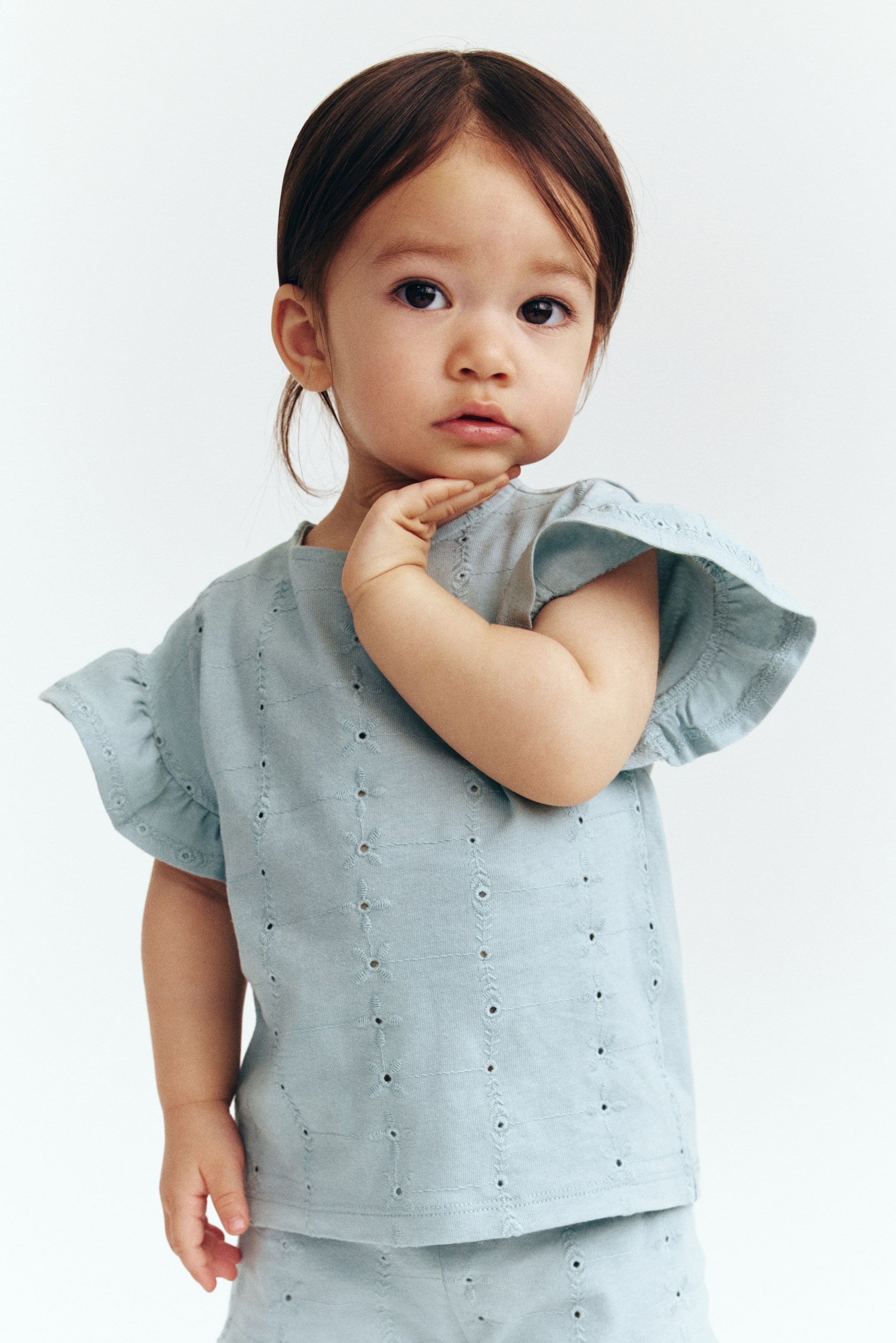 Xem ảnh lớn hơn: Bộ áo và quần short màu xanh nhạt cho trẻ em có thêu lỗ khắp nơi, với tay áo ngắn có bèo trên áo. Quần short đi kèm có thể nhìn thấy bên dưới, cũng hiển thị họa tiết thêu lỗ, và bộ đồ được trình bày từ góc nhìn phía trước.