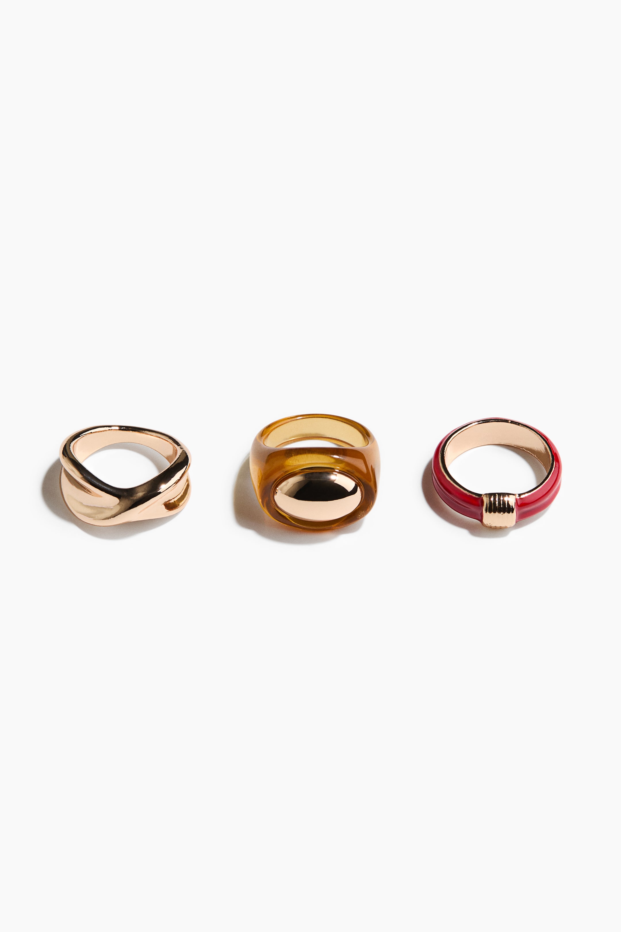 View larger image: Three chunky rings are displaying their designs, with two in metal and one in resin. The first metal ring features a gold-toned crossover band, the resin ring showcases a translucent amber band with a gold-toned oval insert, and the second metal ring presents a red enamel band with a ribbed gold-toned detail.