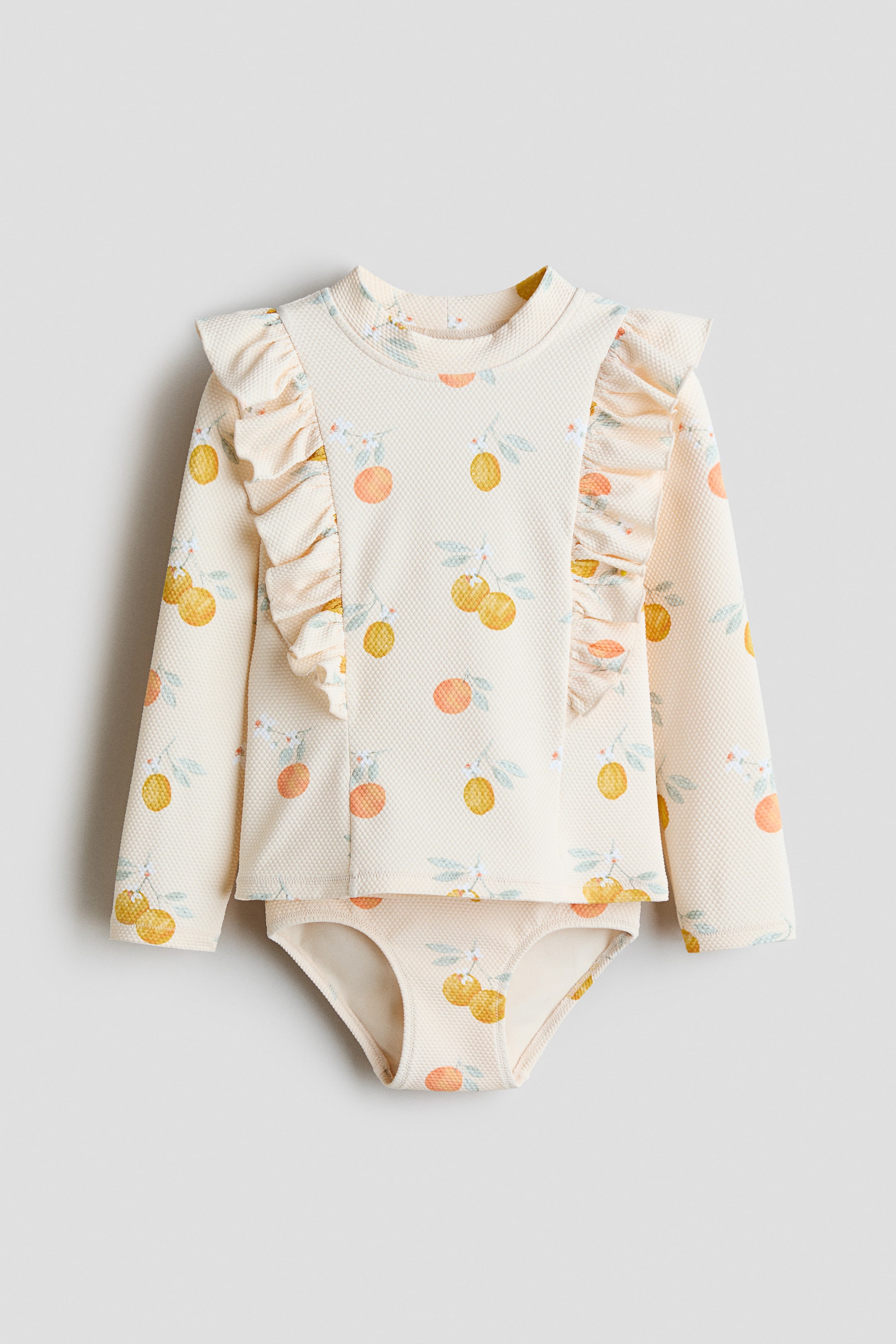 View larger image: An H&M Adorables patterned swim set, shown from the front, features a long-sleeved top with a small stand-up collar and flounces extending from the front over the shoulders to the back. The textured cream fabric of both the top and the visible bikini bottoms displays a vibrant pattern of yellow and orange citrus fruits and green leaves.
