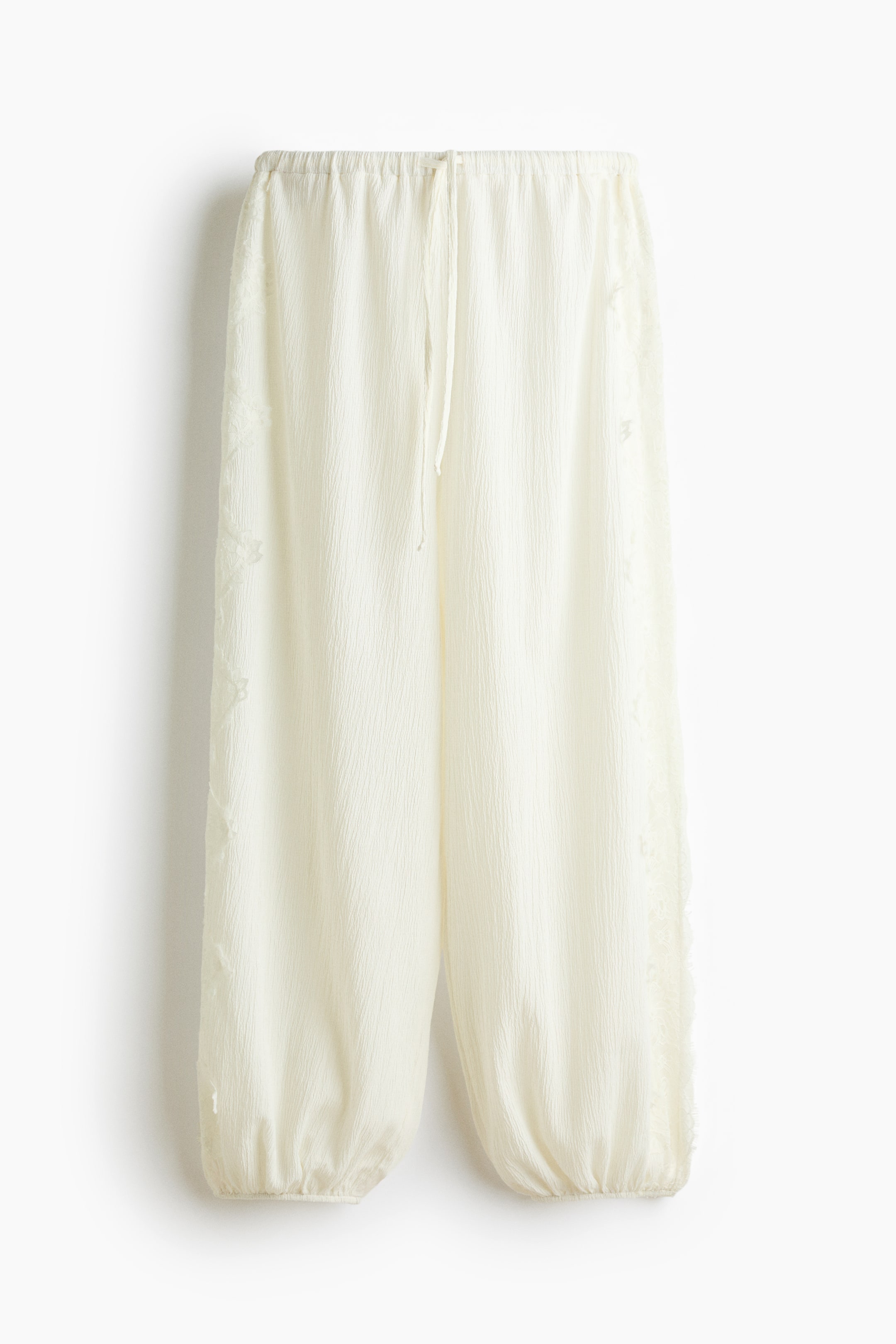 View larger image: Cream-colored pants featuring an elasticated drawstring waistband, a textured, crinkled fabric, and wide legs gathering at elasticated cuffs, with a subtle lace panel detailing the side seams.