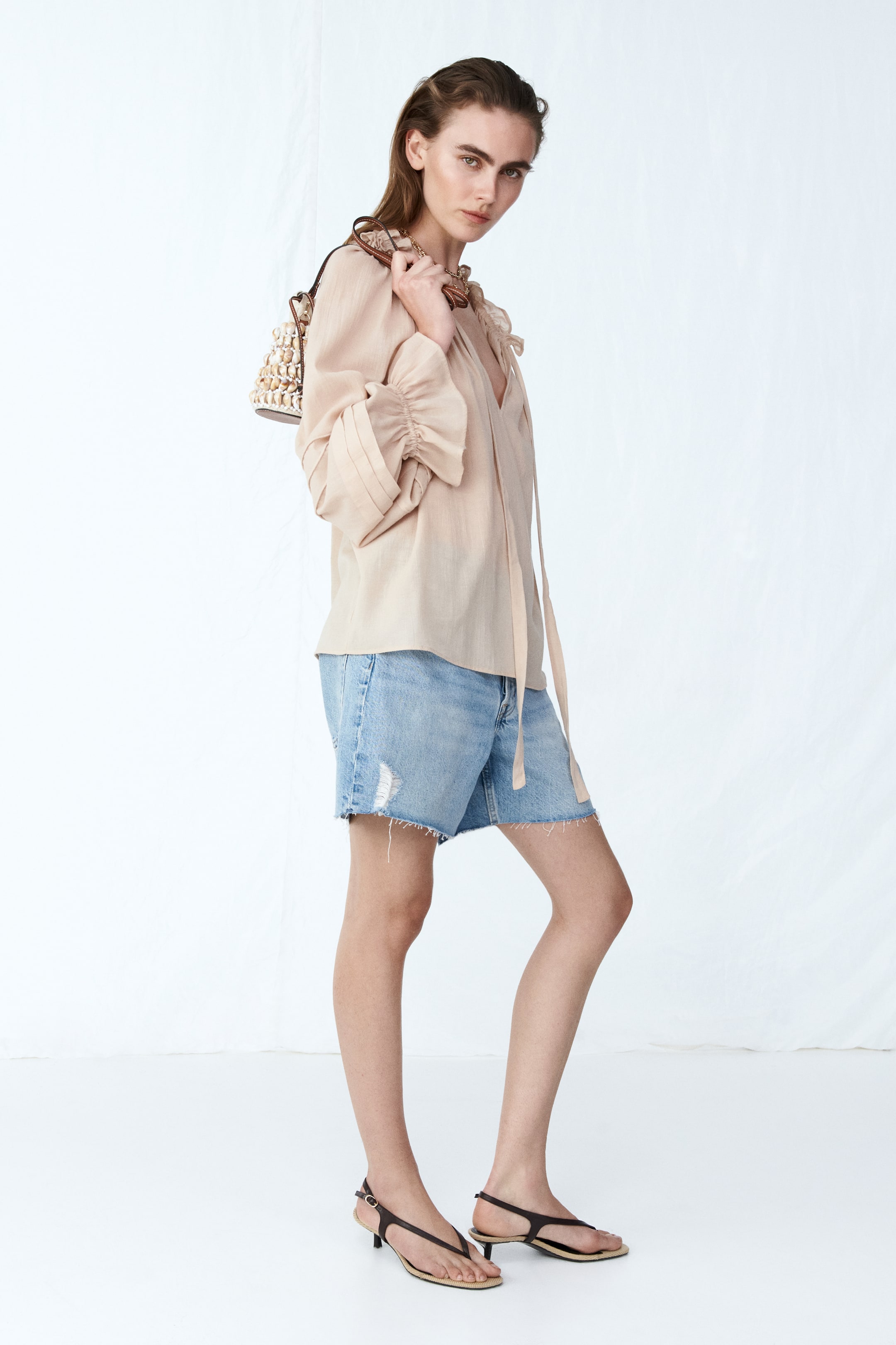 View larger image: A light beige blouse showcases a gathered V-neckline with long ties and voluminous sleeves. The sleeves boast gathered wide cuffs and are styled pushed up, enhancing the blouse's loose, flowing fit as it is worn untucked over denim shorts.