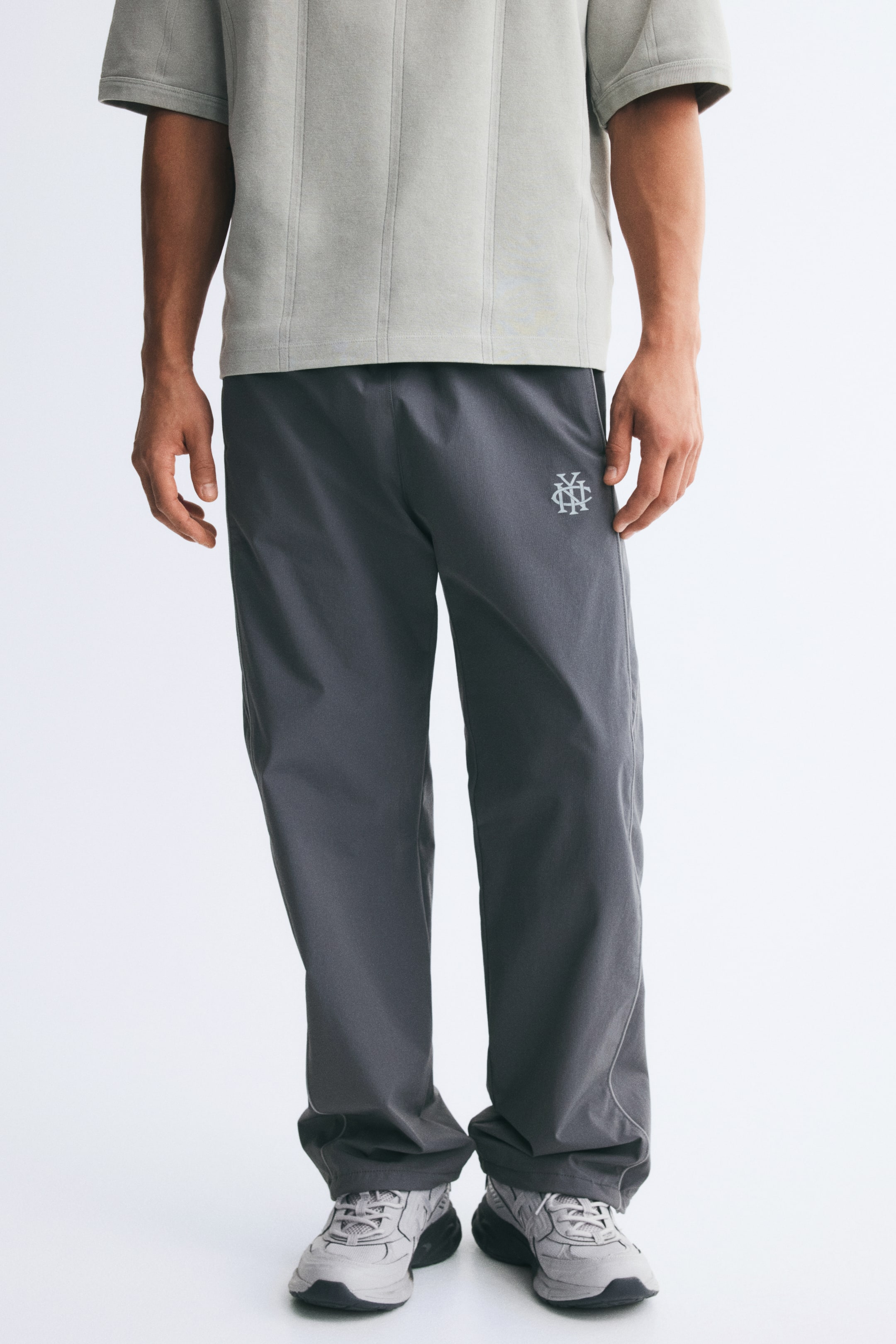 View larger image: The model is wearing track pants in a dark gray ripstop weave, featuring a relaxed fit, a small white embroidered monogram on the upper left thigh, and subtle piping along the side seams. The pants are styled with a loose light gray T-shirt and gray athletic sneakers, with the hems gently gathering at the ankles.