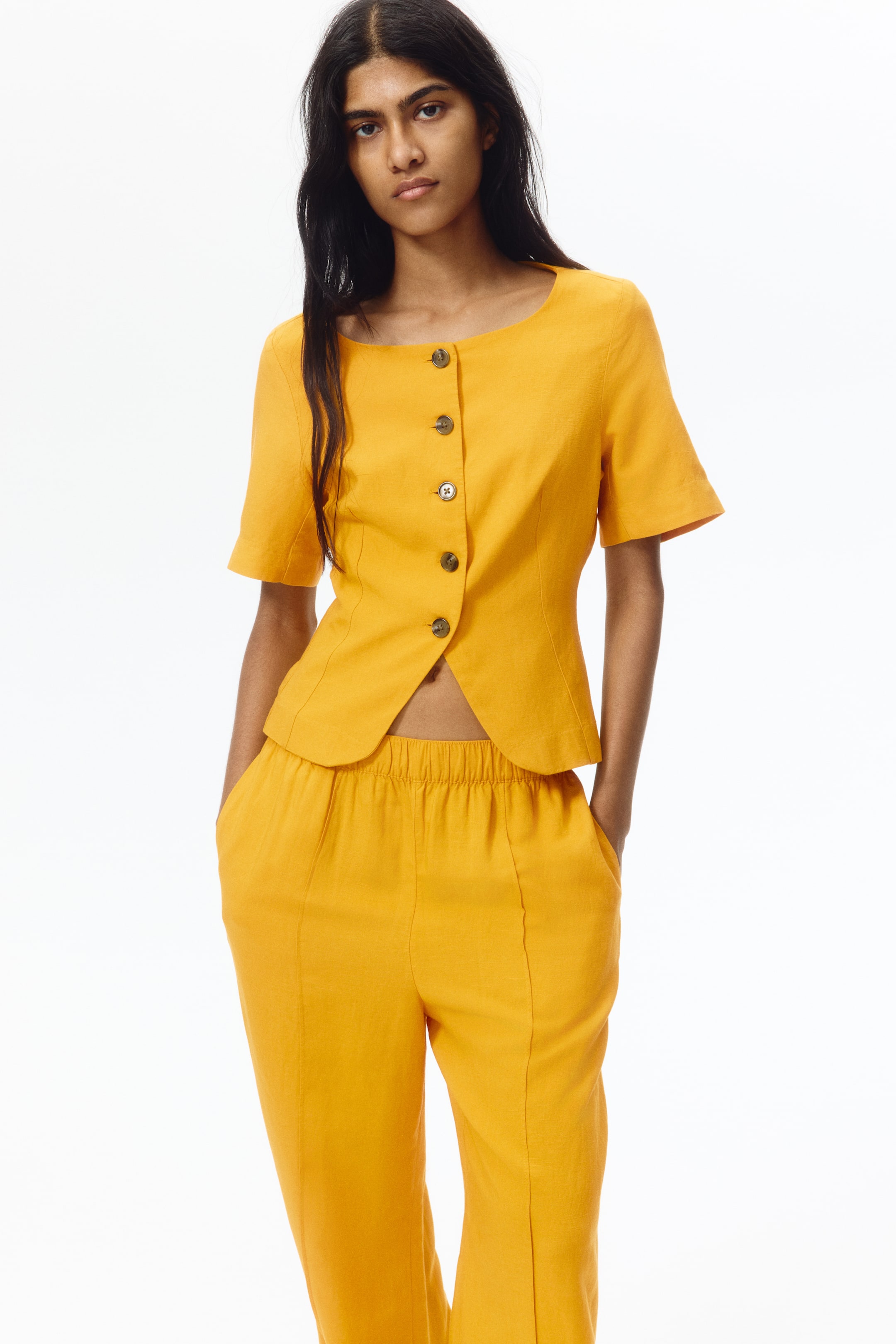 View larger image: A yellow two-piece outfit featuring a cropped, short-sleeved top with a scoop neckline and five dark front buttons. The matching pants display an elasticated waistband, side pockets, and front seams, with the product facing forward.