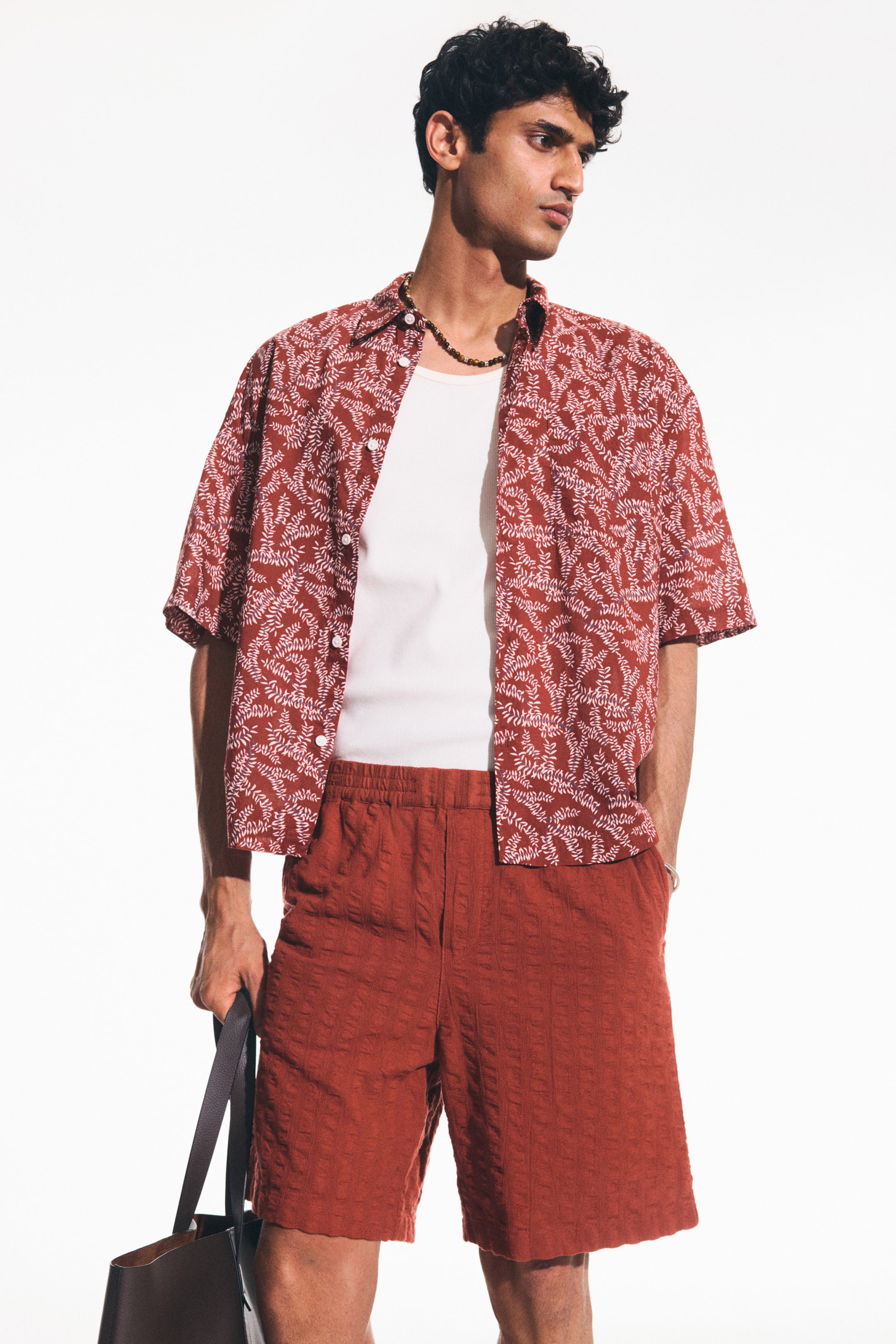 View larger image: The model is wearing reddish-brown shorts crafted from a textured cotton weave, featuring a covered elasticated waist and diagonal side pockets. With one hand casually placed in a pocket, the shorts exhibit a relaxed fit.