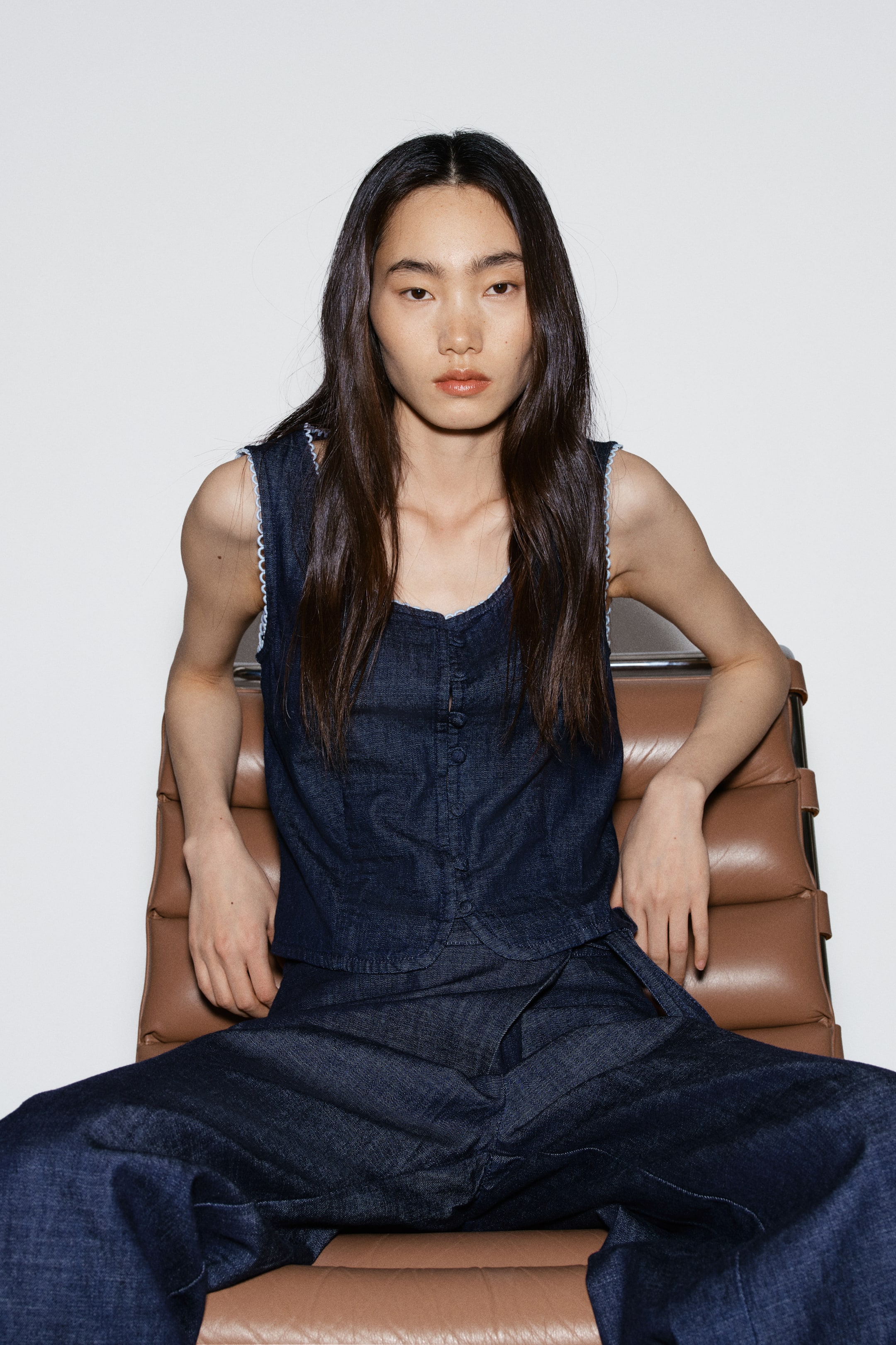 이미지 확대 보기: 어두운 데님 민소매 탑은 V자 네크라인과 버튼 프론트 클로저, 암홀과 네크라인을 따라 흰색 스캘럽 트림이 특징입니다. 어두운 데님 와이드 레그 트라우저가 세트로 구성되어 있으며, 왼쪽 엉덩이에 랩 스타일 클로저가 눈에 띕니다.