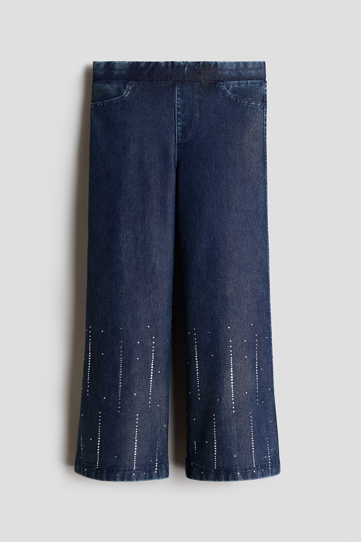 These dark blue, denim-look jersey jeggings are shown from the front, featuring an elasticated waistband, a mock fly, and decorative scoop pockets. The straight-cut legs display vertical patterns of small, silver-coloured rhinestones extending upward from the bottom hems.