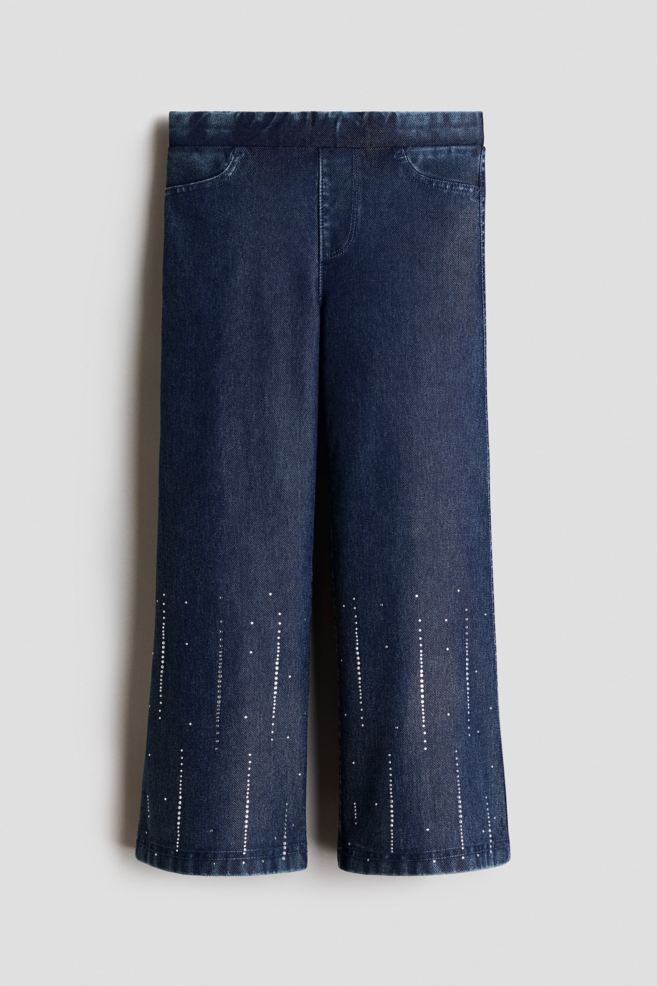 Voir une image plus grande: Ces jeggings en jersey d’aspect denim, de couleur bleu foncé, sont présentés de face et comportent une taille élastiquée, une fausse braguette et des poches décoratives arrondies. Les jambes à coupe droite présentent des motifs verticaux de petits strass argentés s’étendant vers le haut à partir des ourlets du bas.