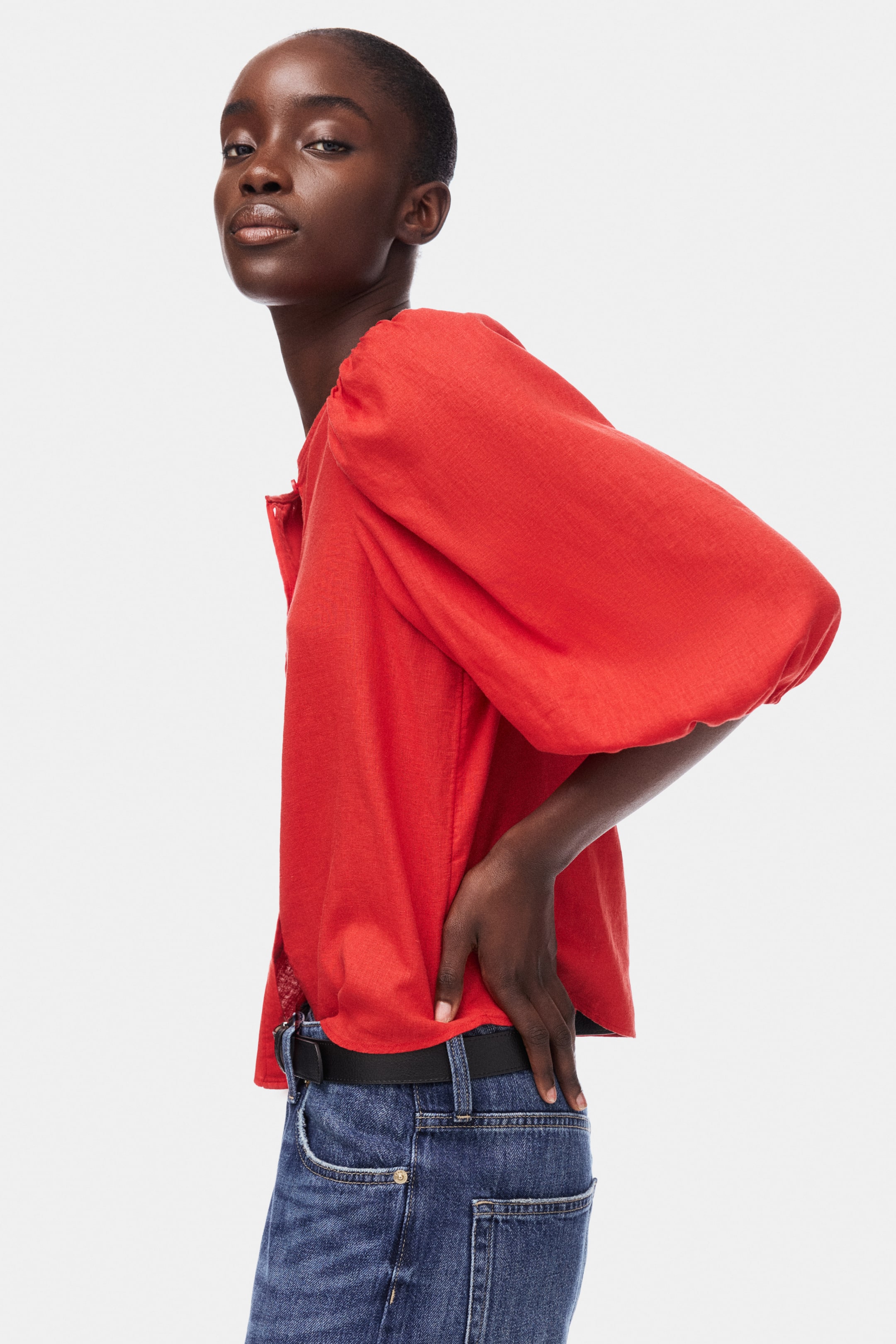 View larger image: The red top shows a subtle woven texture and voluminous three-quarter-length puffed sleeves, gathered at the shoulders and mid-arm. It also features a round neckline with a short button placket and is shown from a side angle, partially tucked into blue jeans.