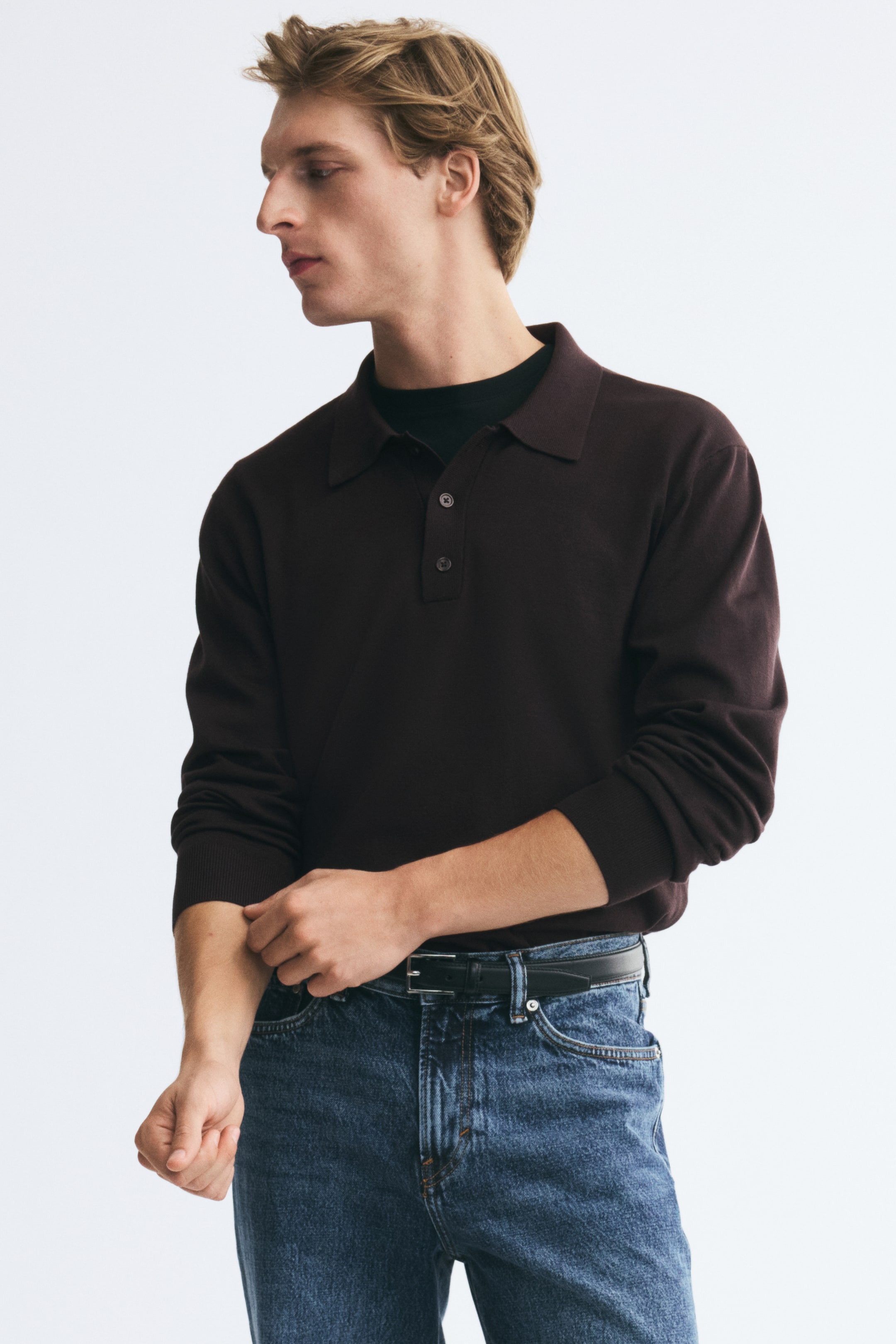 Visualizza immagine più grande: Una polo a maniche lunghe in maglia fine di colore marrone scuro è indossata dal modello, che sta sistemando il polsino a coste di una manica. La polo presenta un classico colletto e una chiusura con bottoni, ed è indossata sopra una T-shirt nera girocollo e infilata in jeans blu con una cintura nera.