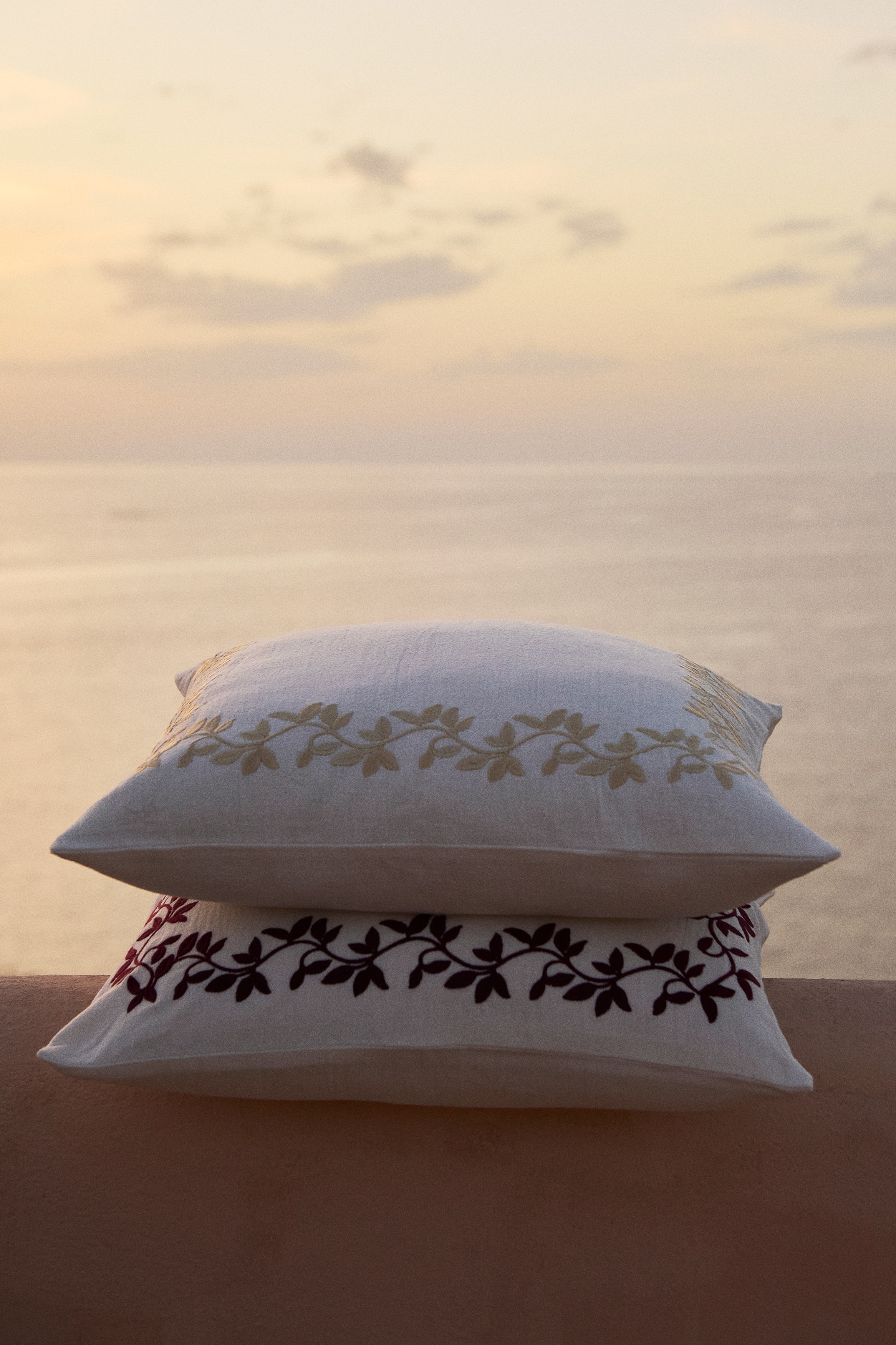 A square white scatter cushion is shown from the front, featuring a textured woven cover with a delicate, light beige embroidered vine and leaf border framing the centre.