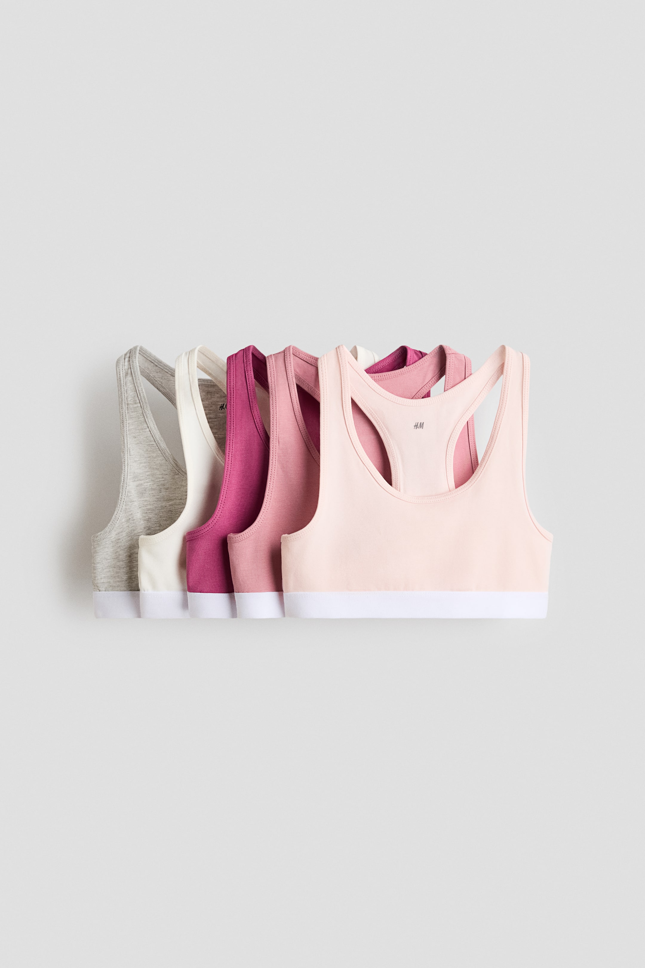 View larger image: Five soft cotton-jersey tops are shown from the front, each featuring a racerback design and a white elasticated hem. The tops are presented in a range of colours, including light pink, dusty pink, fuchsia, off‑white and light grey.