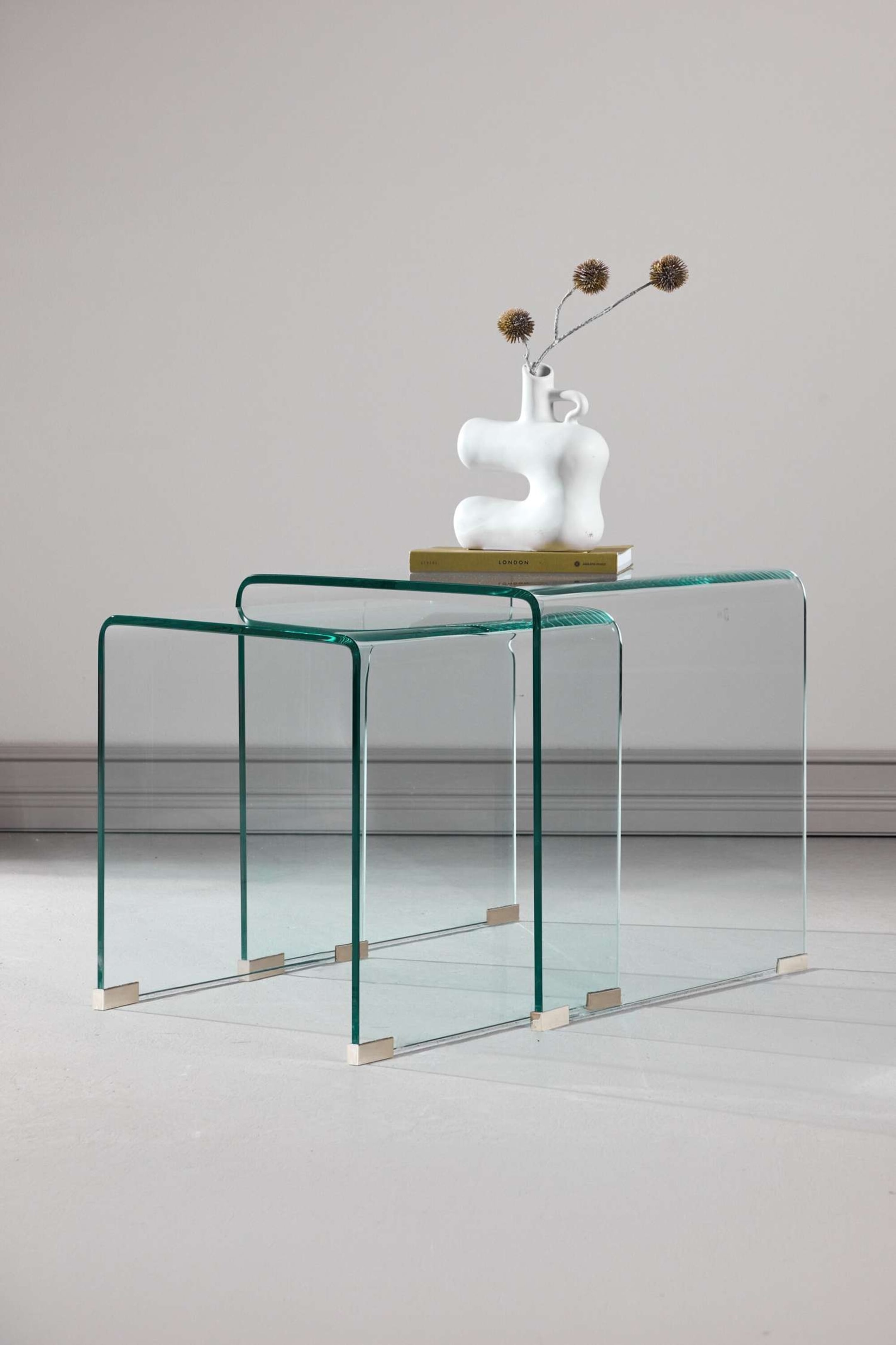 Größeres Bild ansehen: Ein Set aus zwei klaren Glas-Nesttischen ist leicht nach rechts gewinkelt dargestellt. Die Tische zeichnen sich durch gebogene U-förmige Glasstrukturen aus, wobei der kleinere Tisch unter den größeren passt und kleine, hellfarbene Gleiter an den unteren Ecken sichtbar sind. Eine weiße abstrakte Vase auf einem Buch ruht auf dem größeren Tisch.