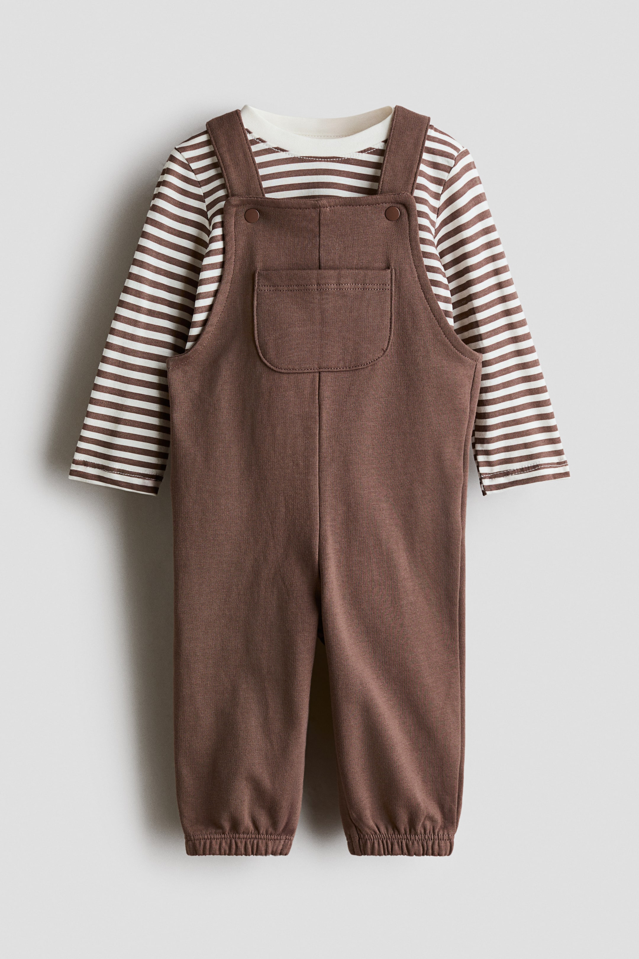 View larger image: A two-piece baby outfit is shown facing forward, featuring solid brown overalls layered over a long-sleeved crewneck t-shirt with brown and white horizontal stripes. The overalls feature snap-button shoulder straps, a central chest patch pocket, and elasticated ankle cuffs.