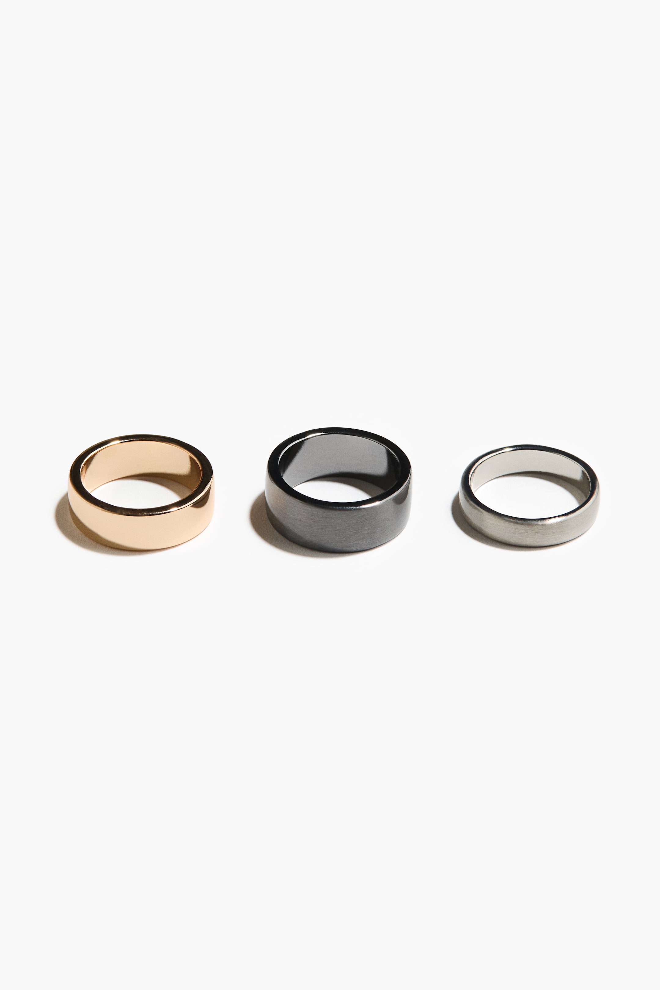 View larger image: Three rings are showcased in a horizontal arrangement, each displaying a unique finish and width. On the left, a broad, polished gold-toned band is featured. In the center, a wide, brushed dark grey band is highlighted, while on the right, a narrower, brushed silver-toned band is presented.