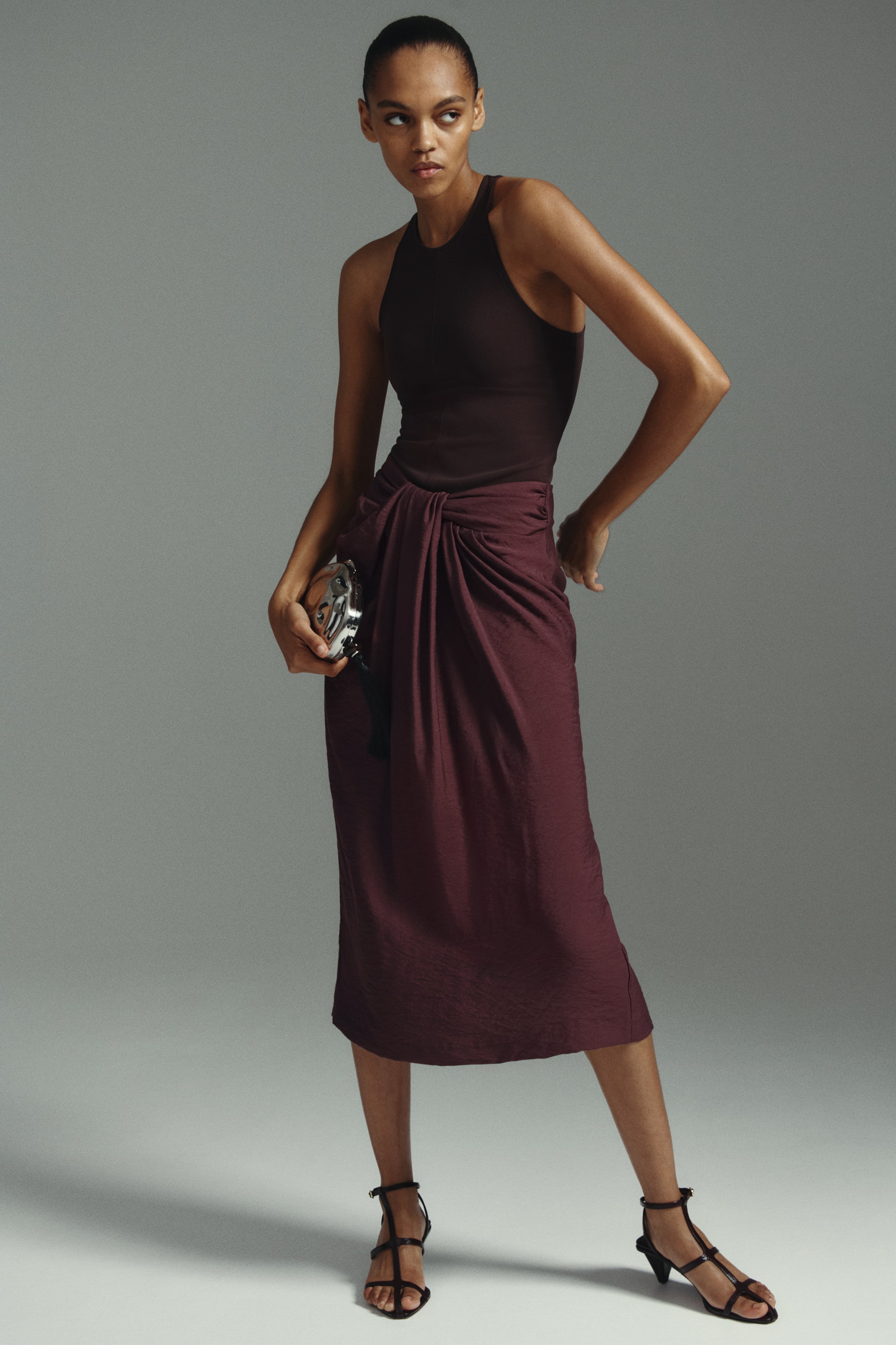 Größeres Bild anzeigen: Ein dunkelbraunes, ärmelloses Top mit hohem Kragen wird mit einem bordeauxroten Midirock getragen, der ein drapiertes, geknotetes Detail an der vorderen Taille präsentiert. Schwarze Riemchensandalen mit niedrigen Absätzen vervollständigen das Ensemble.