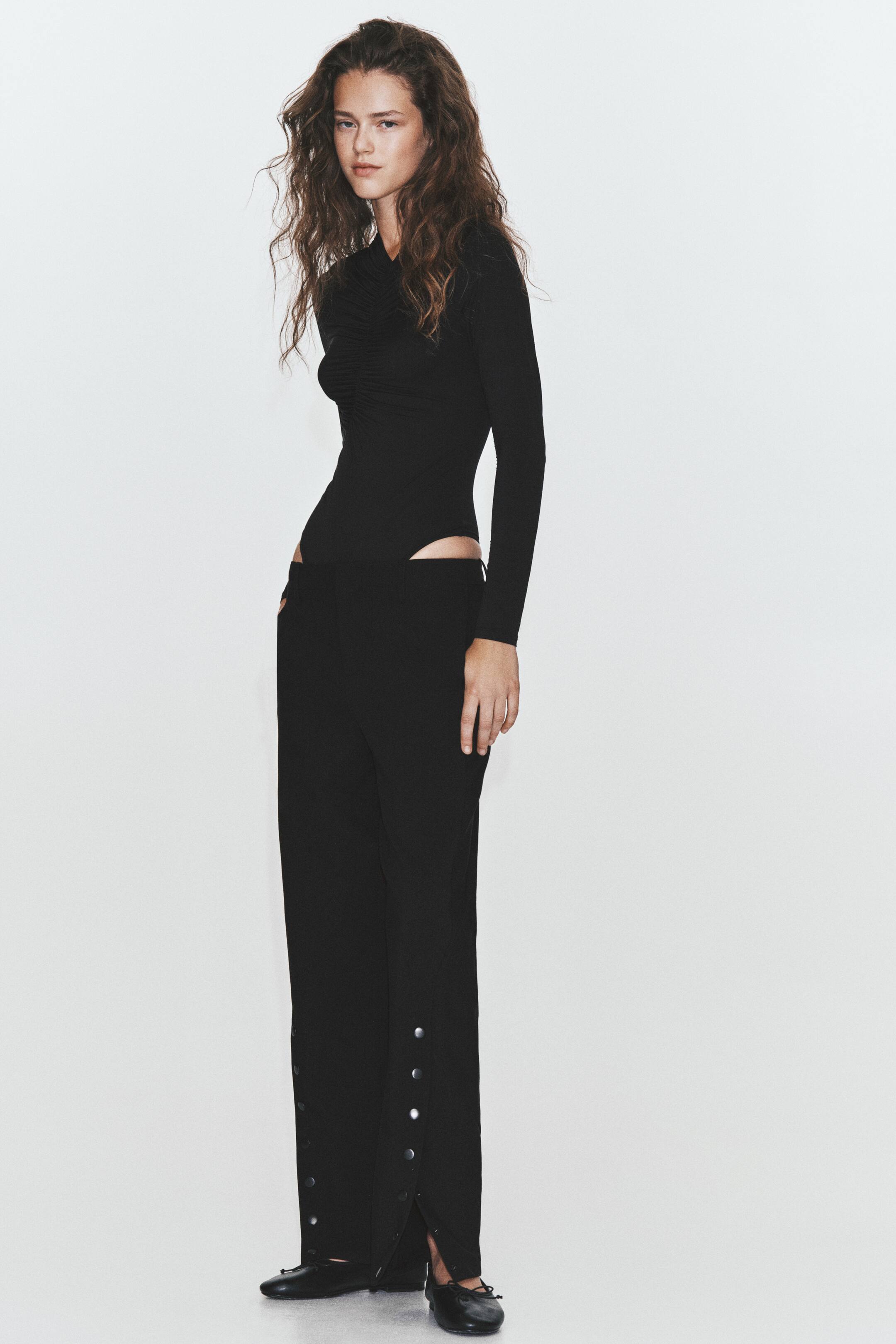 Ampliar la imagen: Pantalones negros de sarga con silueta de pierna ancha y aberturas altas con broches de presión plateados en el dobladillo, llevados por la modelo. La modelo está de pie y combina los pantalones con un body negro de manga larga y zapatos negros planos.