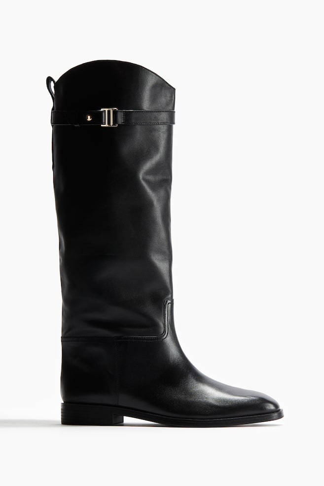 A black soft leather boot is presented from a side angle, featuring a rounded toe and a knee-high shaft with a slanted top. It is detailed with a strap and a gold-toned metal buckle near the top, a pull loop at the back, and decorative seams. - 1