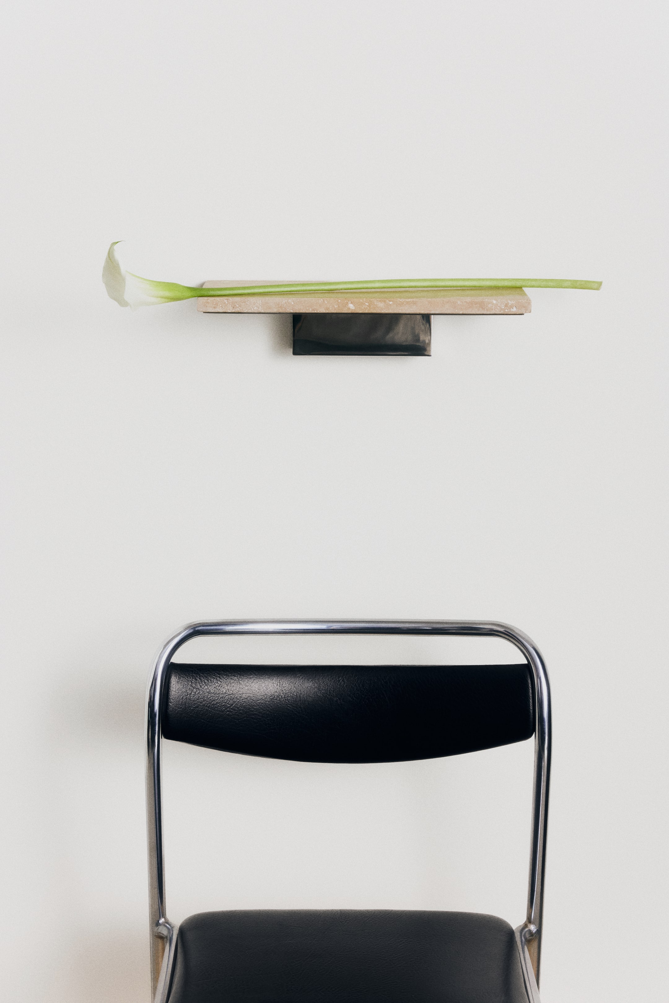 View larger image: A wall shelf featuring a rectangular travertine stone slab supported by a dark stainless steel bracket is shown from the front. A single white calla lily with a long green stem rests horizontally across the shelf surface.
