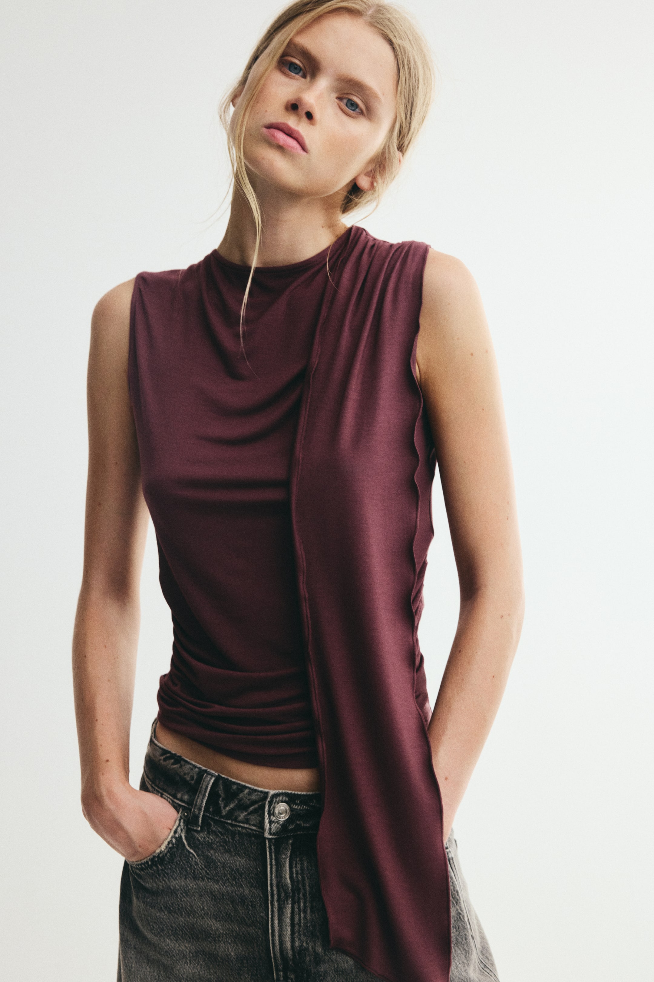 View larger image: A sleeveless, fitted top in dark red stretch viscose jersey is worn by the model. It features a round neckline, with a long panel and gathers cascading from one shoulder and gathers at the opposite waist, creating a ruched effect.