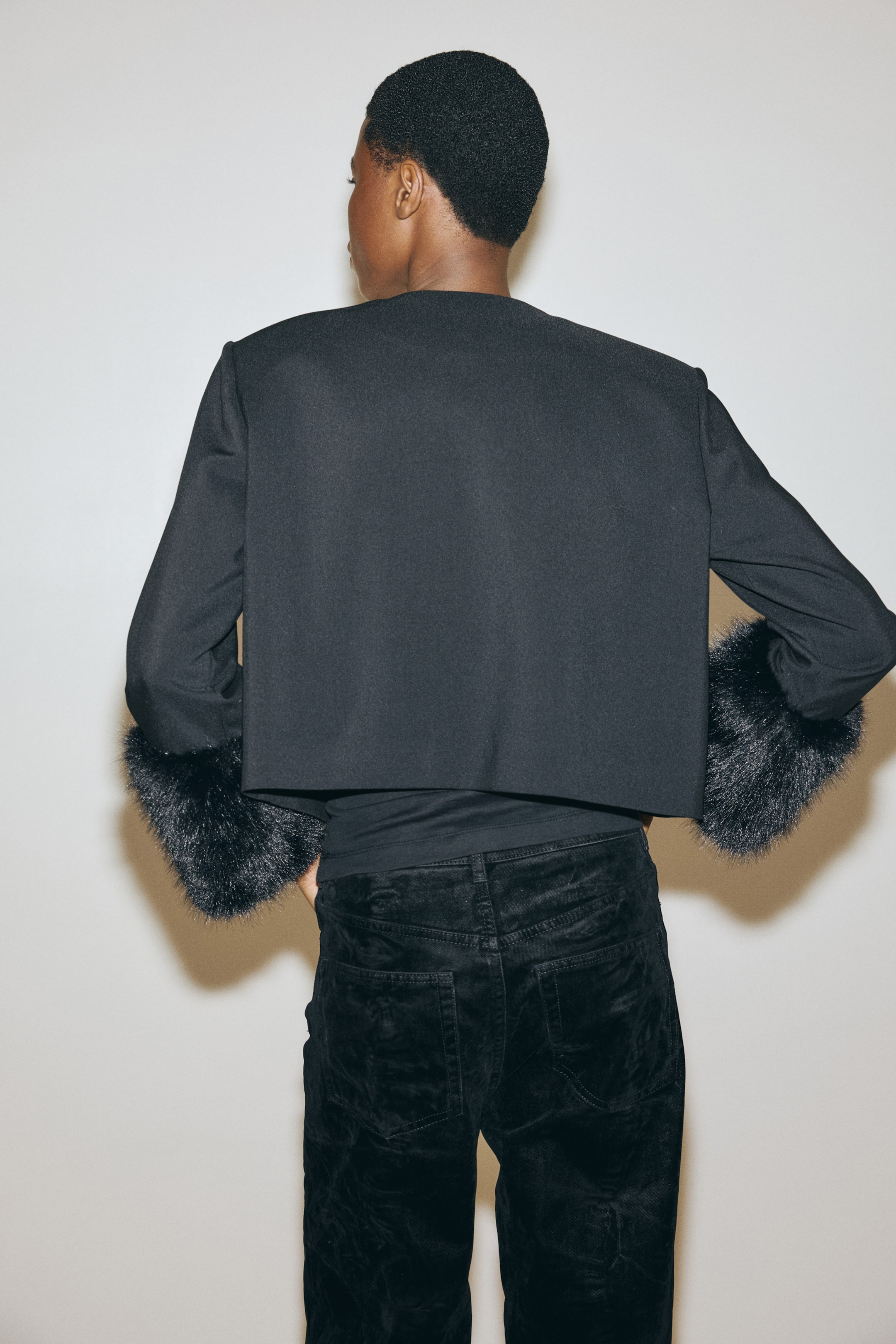 View larger image: A black cropped jacket is being presented from the back, featuring a straight hemline ending above the waist. It is showcasing long sleeves adorned with voluminous black faux fur cuffs.