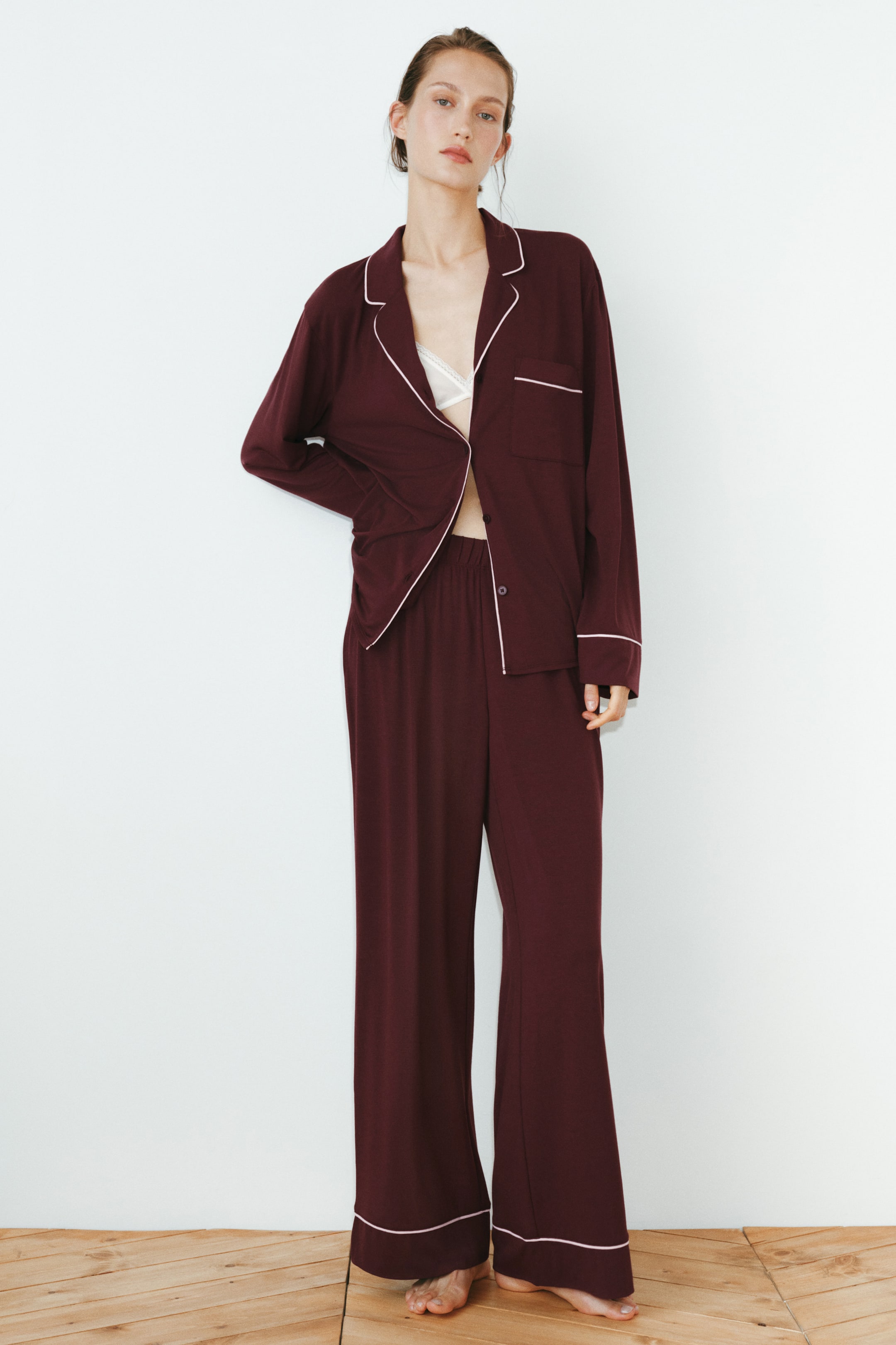 Agrandir l'image: Le mannequin porte un pyjama en jersey rouge foncé. La chemise à manches longues est dotée de revers crantés et de passepoils blancs, et est partiellement ouverte, révélant un bralette blanche en dessous, tandis que le pantalon à jambes larges présente une taille élastiquée et des passepoils blancs aux ourlets.