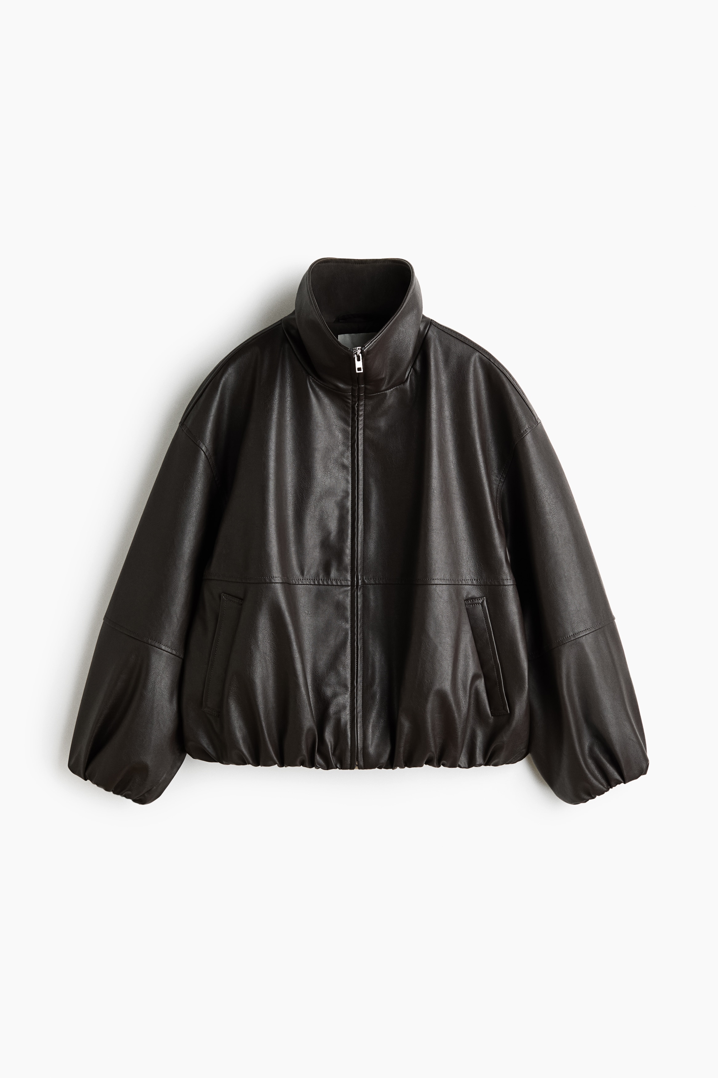 La modelo lleva puesta una chaqueta de corte oversize en tejido oscuro con acabado revestido, combinada con una camisa blanca de cuello. La chaqueta presenta un cuello chimenea alto, hombros caÃ­dos y un bolsillo lateral de ojal en el que la modelo estÃ¡ introduciendo la mano.