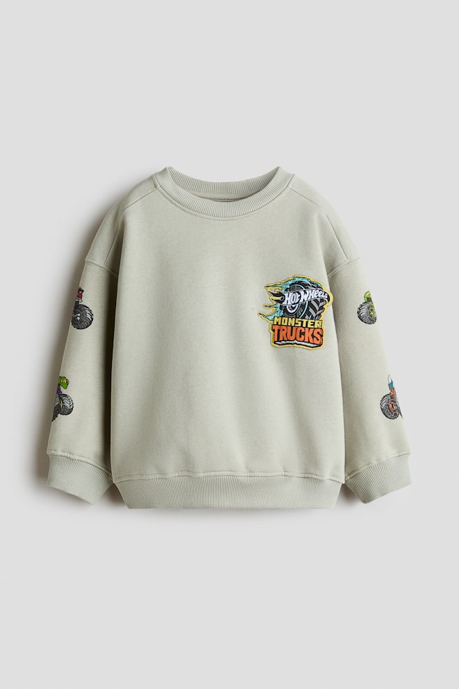 A light sage green sweatshirt is shown from the front, featuring a ribbed crew neck, dropped shoulders, ribbed cuffs and a ribbed hem. The chest displays a "Hot Wheels Monster Trucks" graphic patch, and the sleeves feature small printed monster truck illustrations. - 1