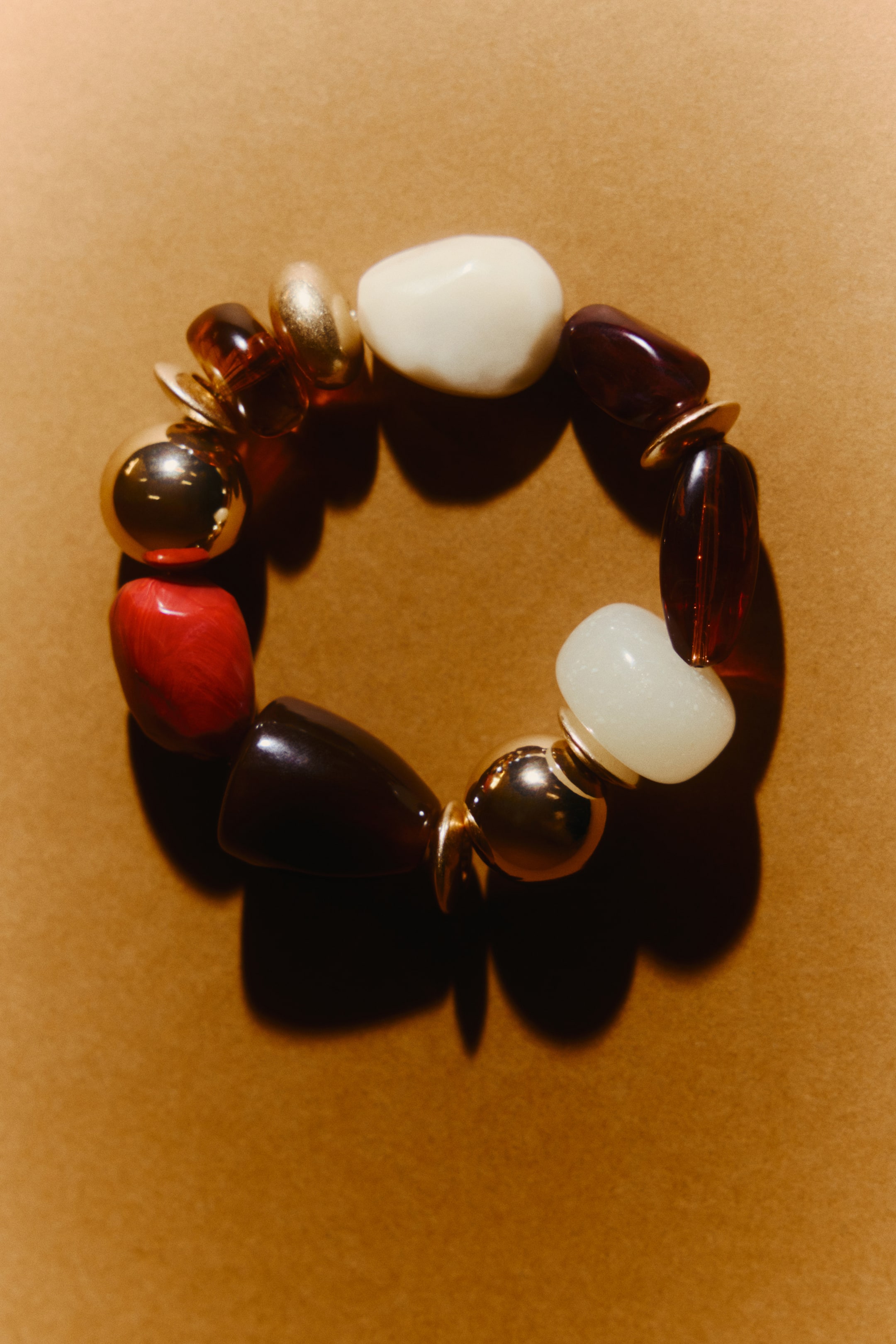 Größeres Bild ansehen: Ein elastisches Armband mit großen Perlen in verschiedenen Formen und Designs liegt flach. Die Perlen sind in opakem Weiß, Rot und Dunkelbraun gehalten und werden durch glänzende goldfarbene Akzente ergänzt.